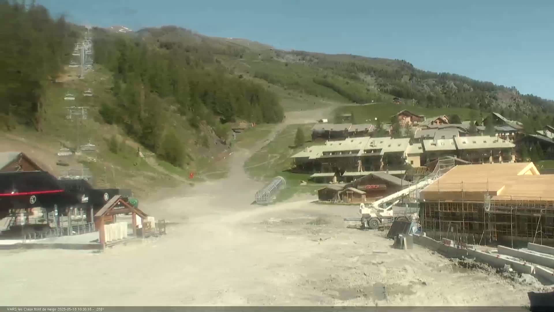 A ski resort village is visible on a sunny day, with a partially constructed building in the foreground and a chairlift extending up a grassy hillside.