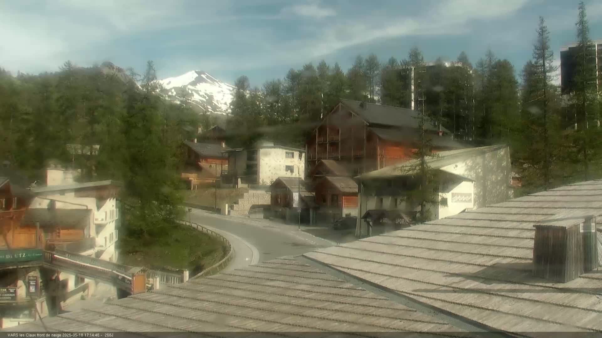 A mountain village nestled in a valley under a partly cloudy sky, with a snow-capped mountain in the background.