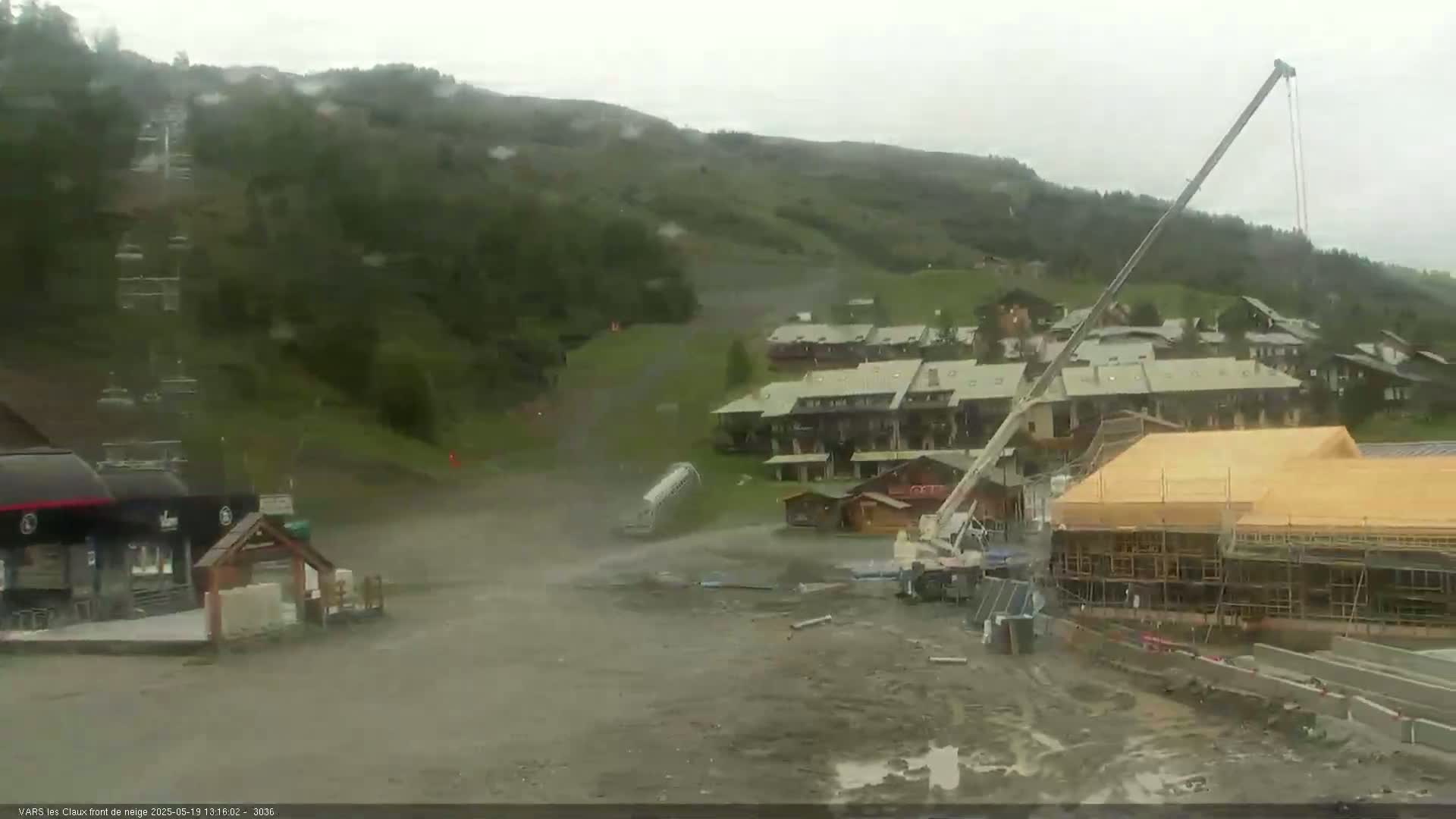 A muddy construction site with a large crane and partially built building is visible near a cluster of buildings on a hillside in rainy weather.