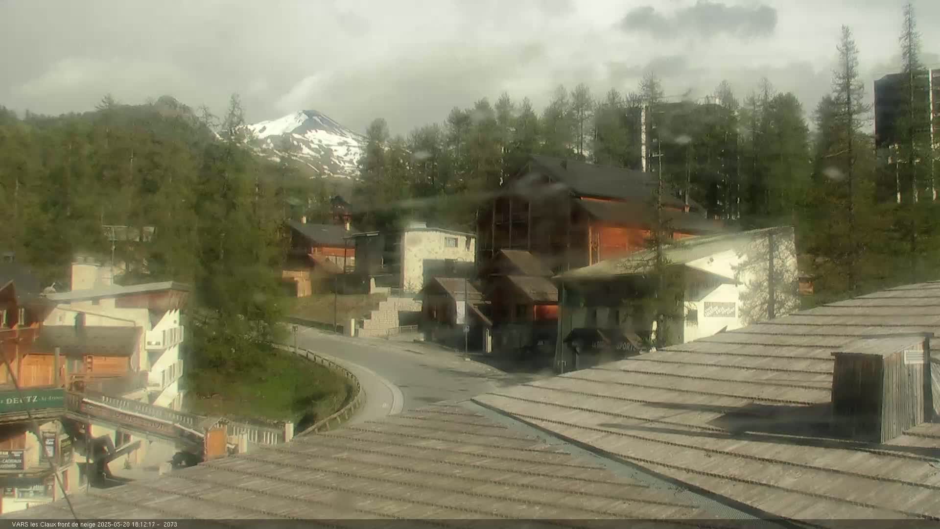A mountain village with wooden buildings is seen on a rainy day, with a snow-capped mountain visible in the background.