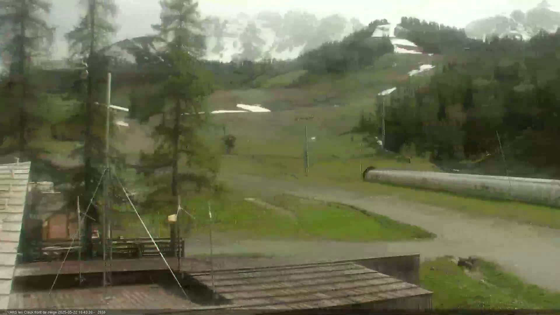 A rainy view of a grassy area with a few trees and buildings, a long pipe, and mountains in the background.