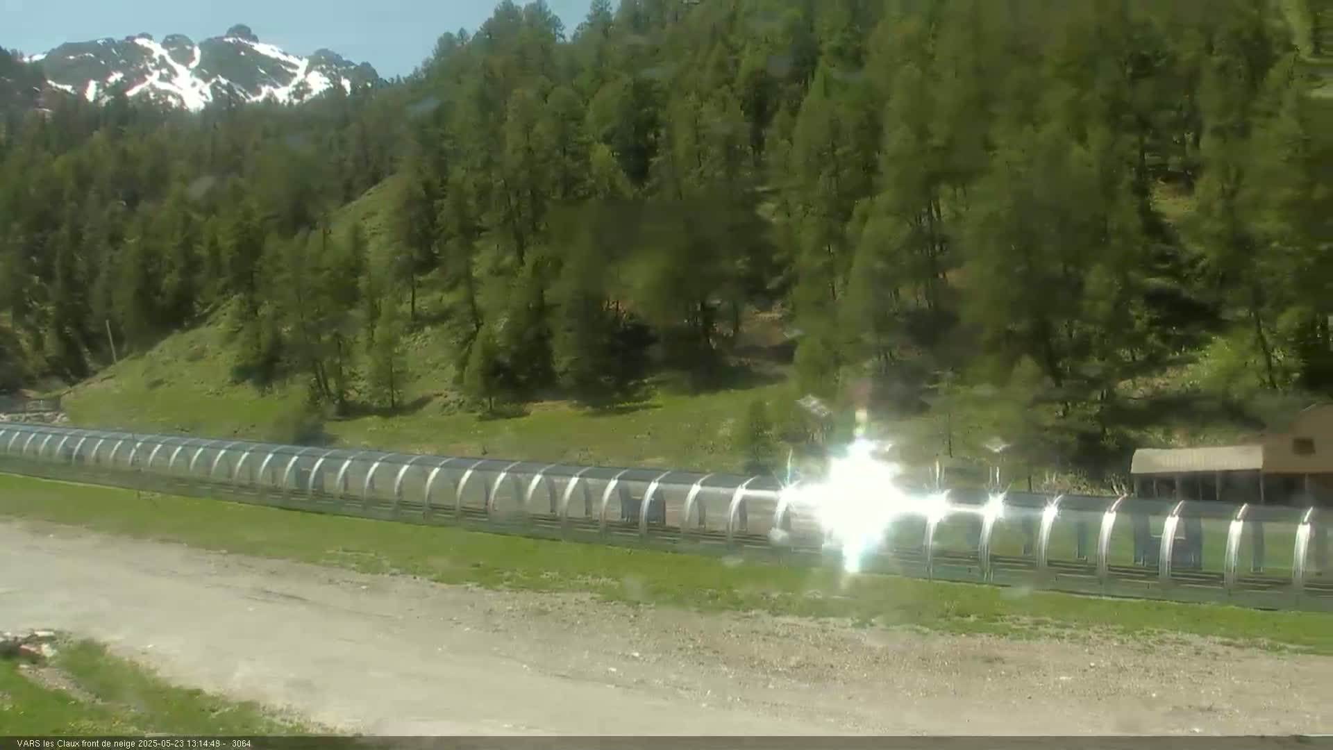 A long, covered walkway stretches across a grassy area beneath a sunny sky, backed by a hillside of evergreen trees and snow-capped mountains in the distance.