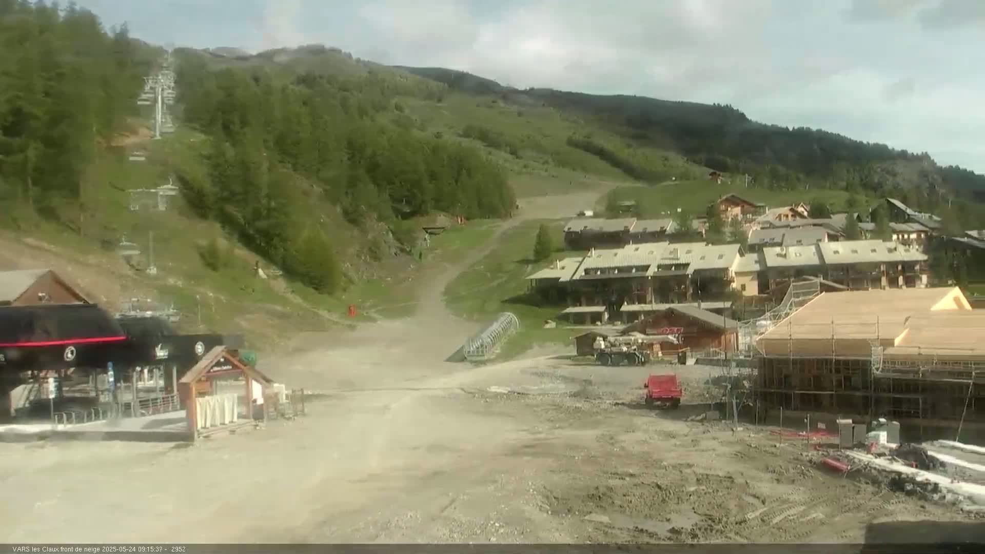 A ski resort in the mountains on a partly cloudy day shows a mostly barren ski slope with a chairlift, buildings, and construction.