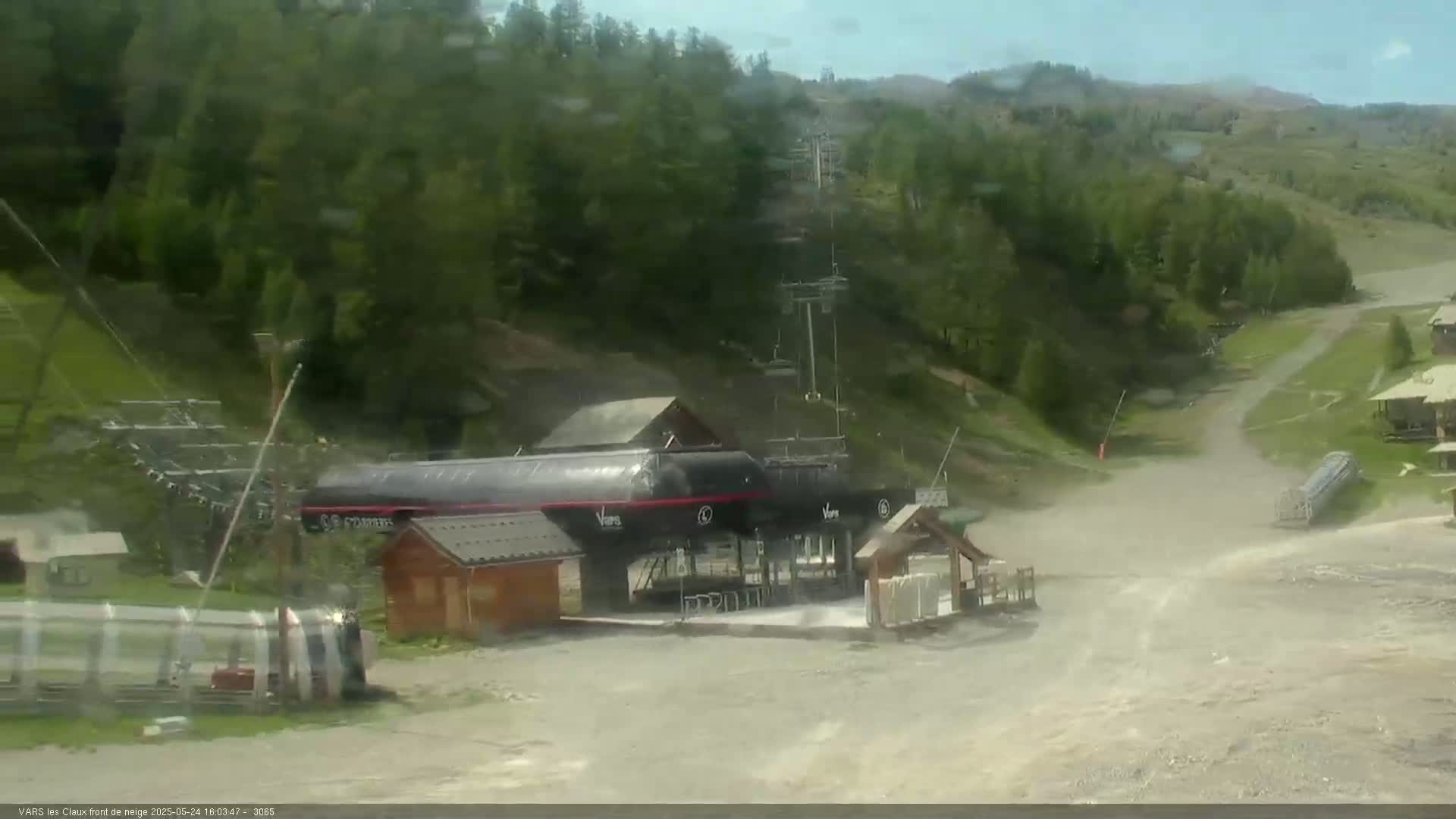 A ski lift base station with several buildings and a chairlift visible in the background, on a partly cloudy day with some dust or light blowing debris on the ground.