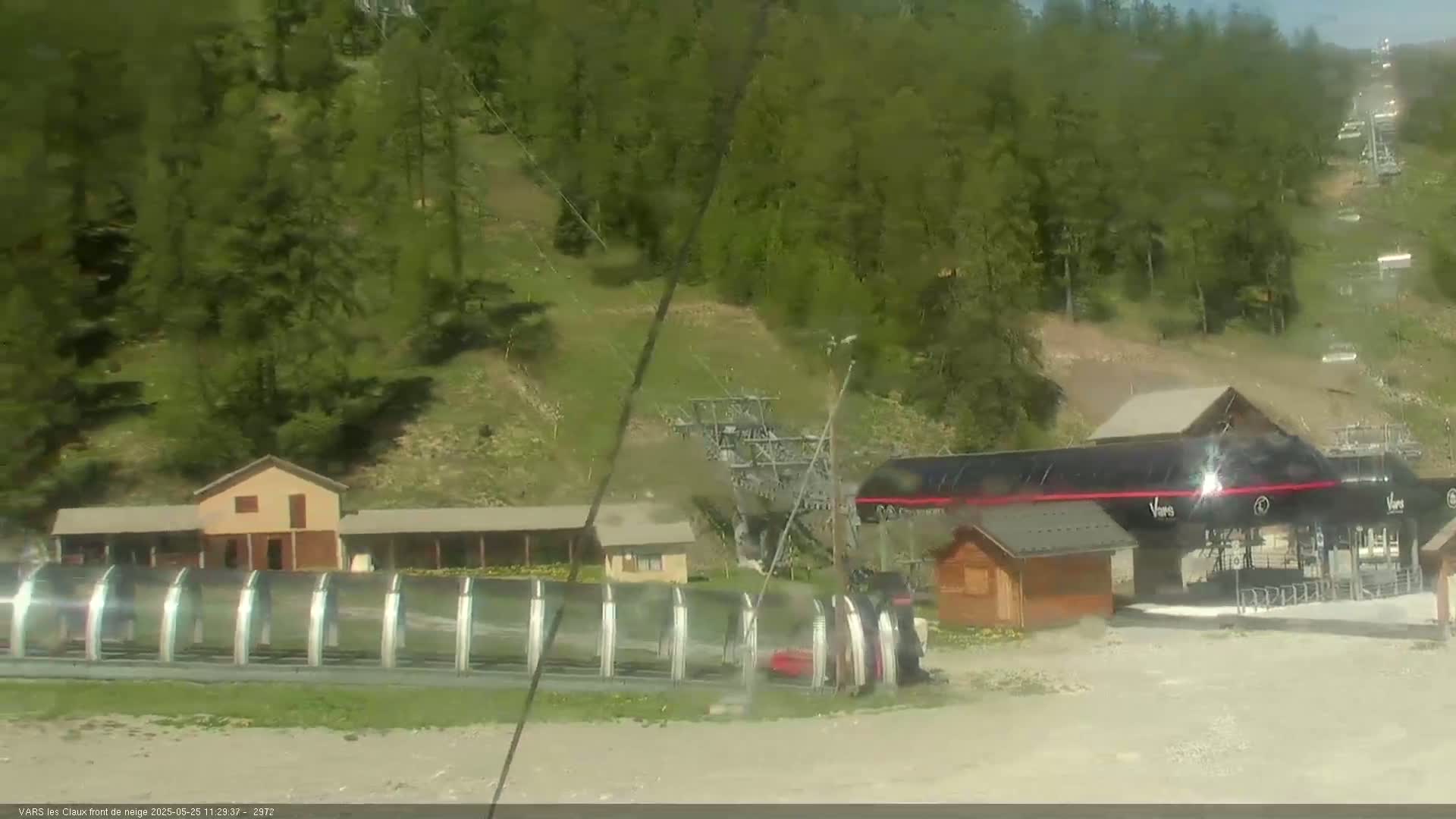 A ski lift base station, with several small buildings and covered loading areas, sits amidst a lush green hillside on a sunny day.