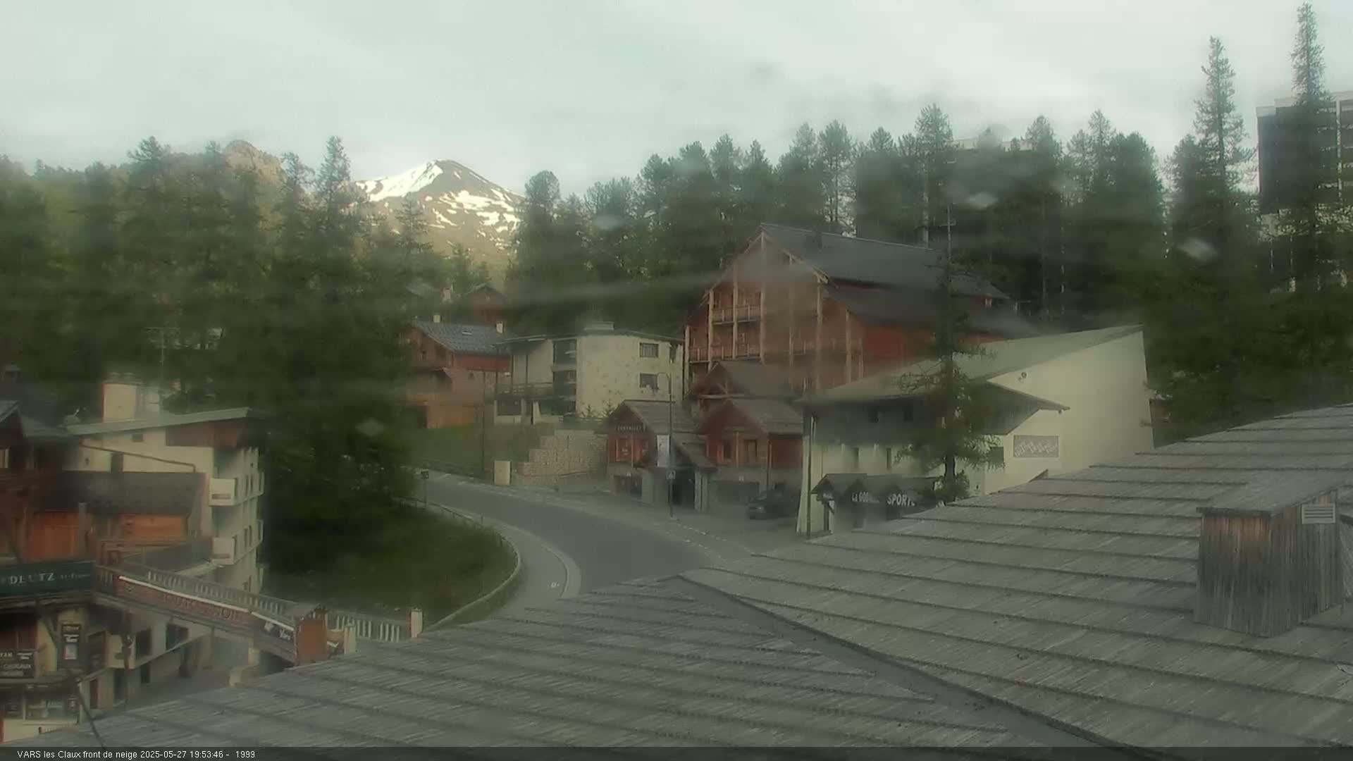 A rain-soaked mountain village scene shows a curving road in front of several chalet-style buildings, with a snow-capped mountain visible in the background.