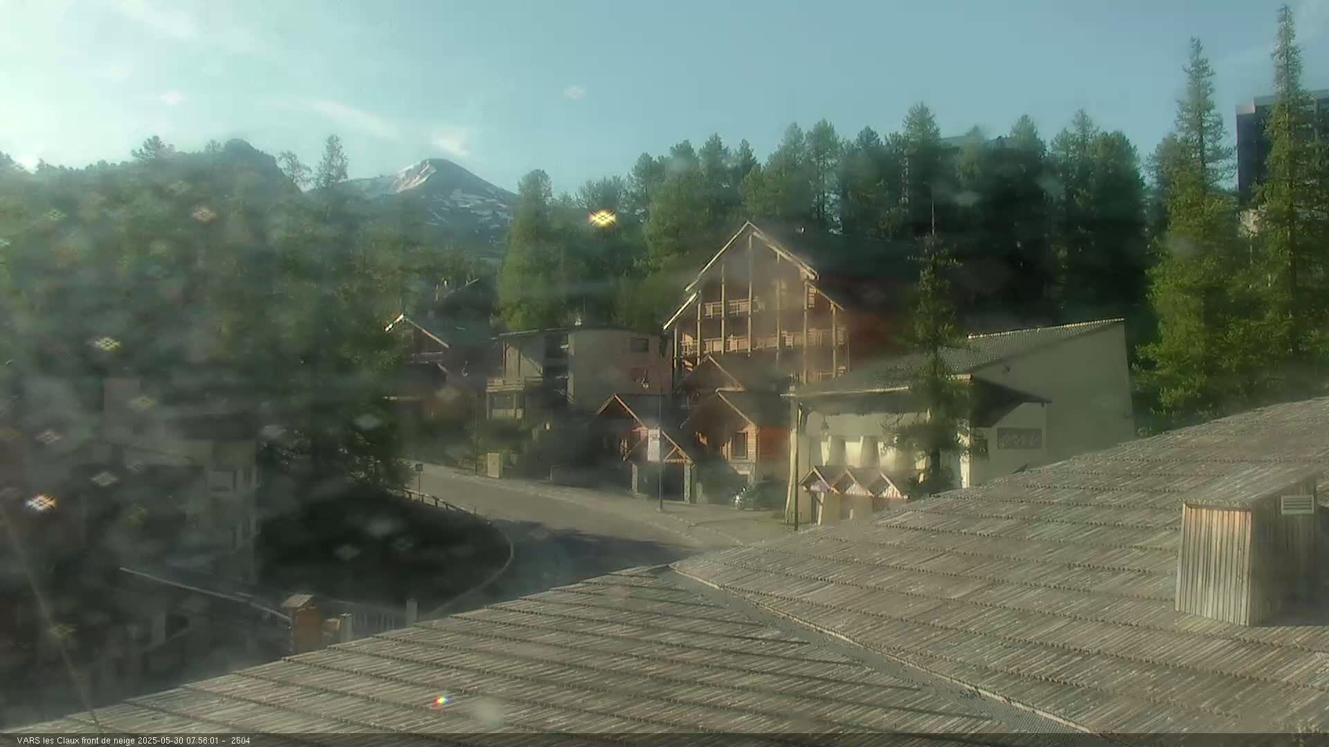 A mountain village nestled among green trees and a snow-capped mountain peak in the distance under a mostly clear sky.