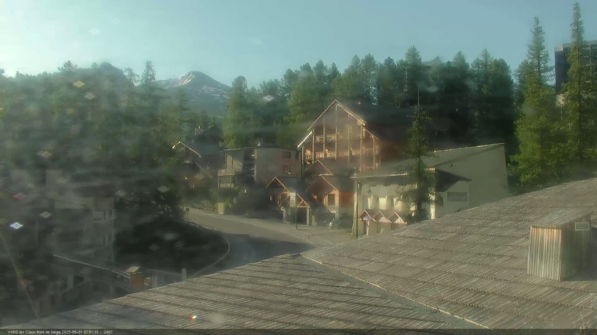 A slightly hazy view of a mountain village with several buildings, including a multi-story wooden structure, nestled among green trees and a snow-capped mountain in the background under a clear sky.