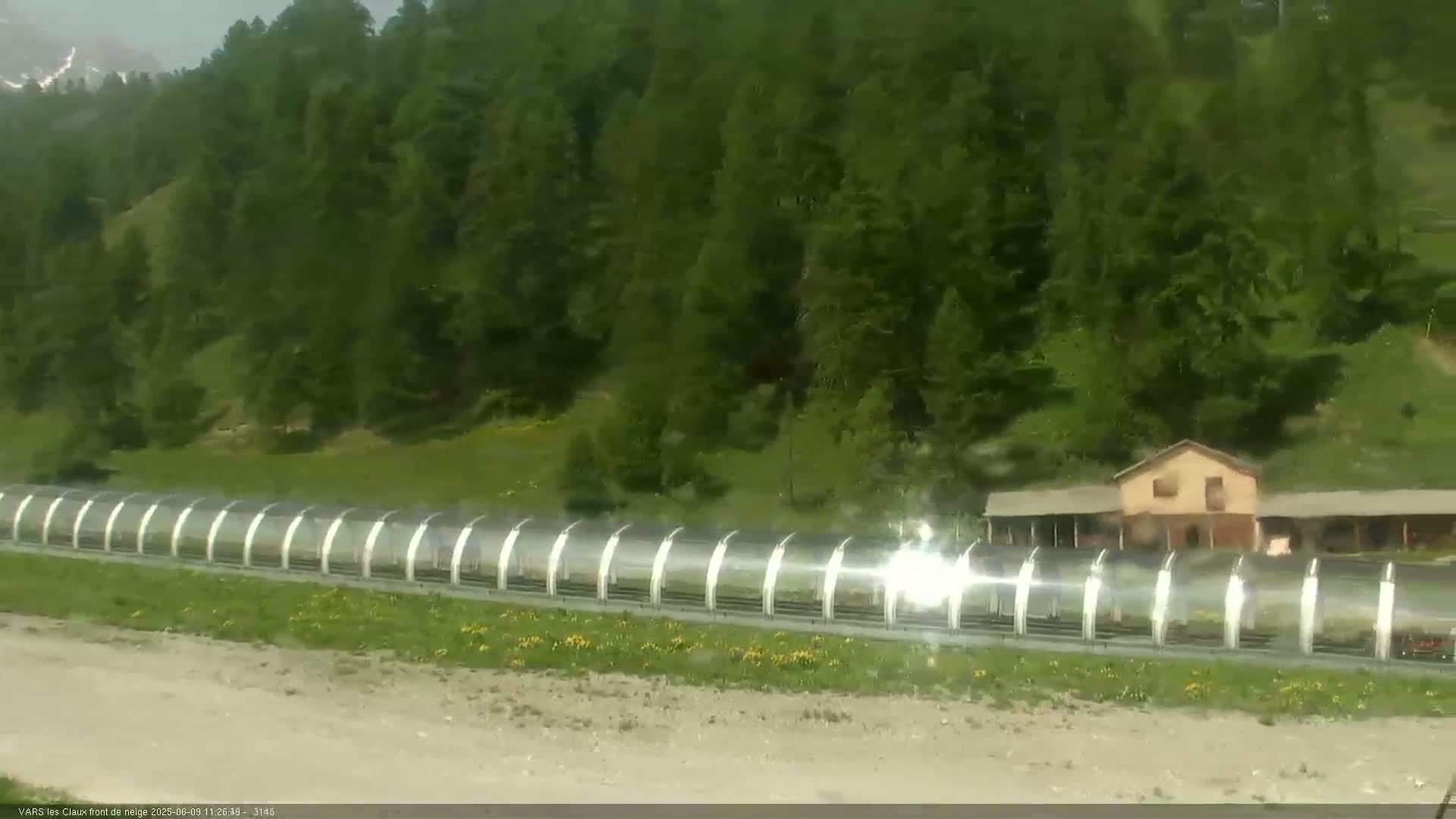 A long, covered walkway extends across a grassy area in front of a building nestled among a hillside of evergreen trees under a bright, sunny sky.