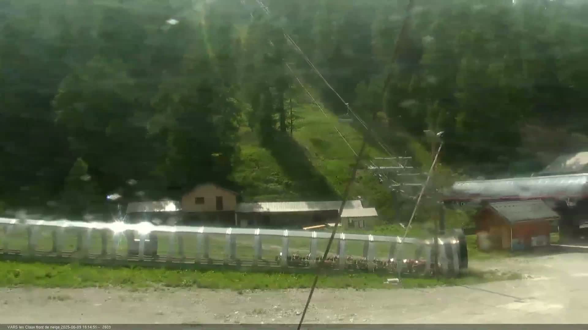 A long row of enclosed chairlifts sits in front of a small building and a large fuel tanker, all situated on a hillside amidst a misty, green forest.