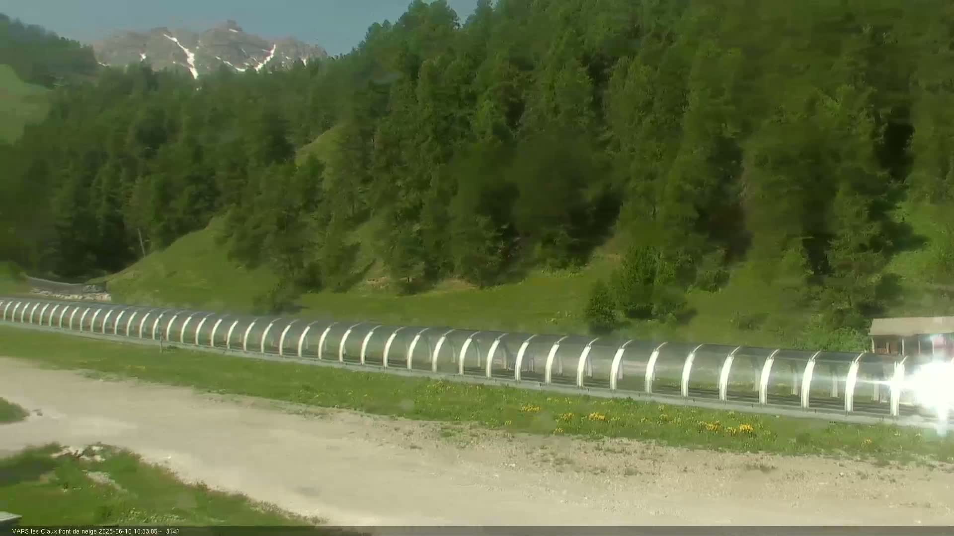 A long, covered walkway stretches across a grassy area below a hillside of green trees and a mountain range under a mostly clear sky.