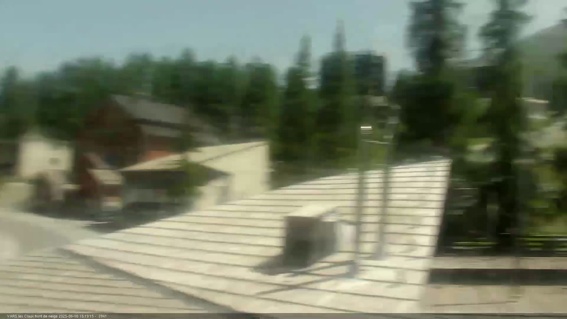 A blurry view from a moving vehicle shows a tan shingled roof, several buildings, and a background of green trees under a sunny sky.