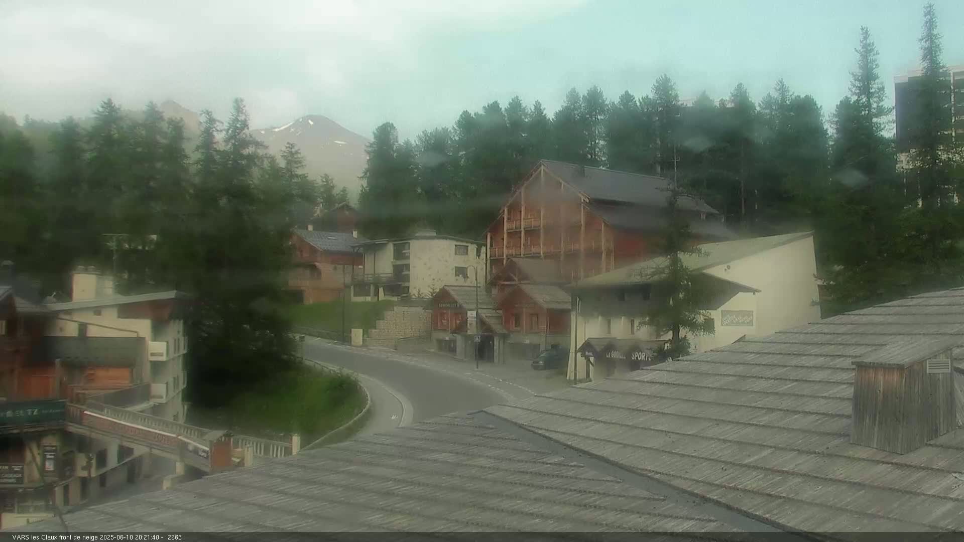 A misty mountain village scene shows several buildings, a curving road, and a mountain range in the background under an overcast sky.