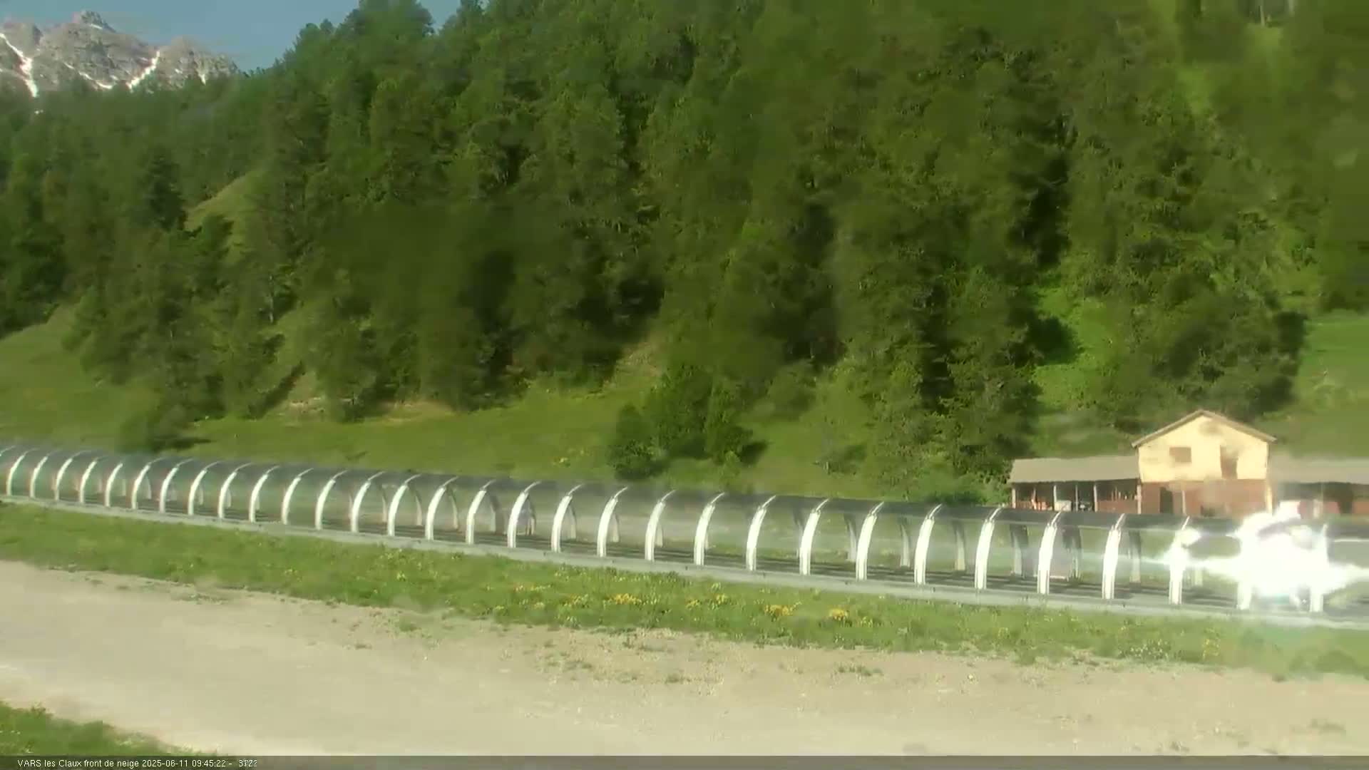 A long covered walkway extends across a grassy area below a hillside of green trees and mountains under a clear sky.