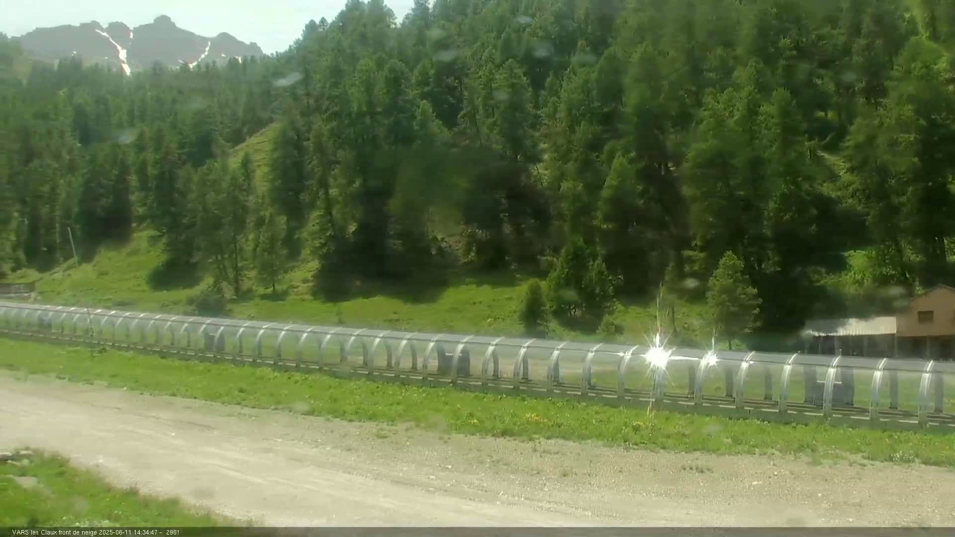 A long, covered structure extends across a grassy area in front of a mountainous forest on a sunny day.