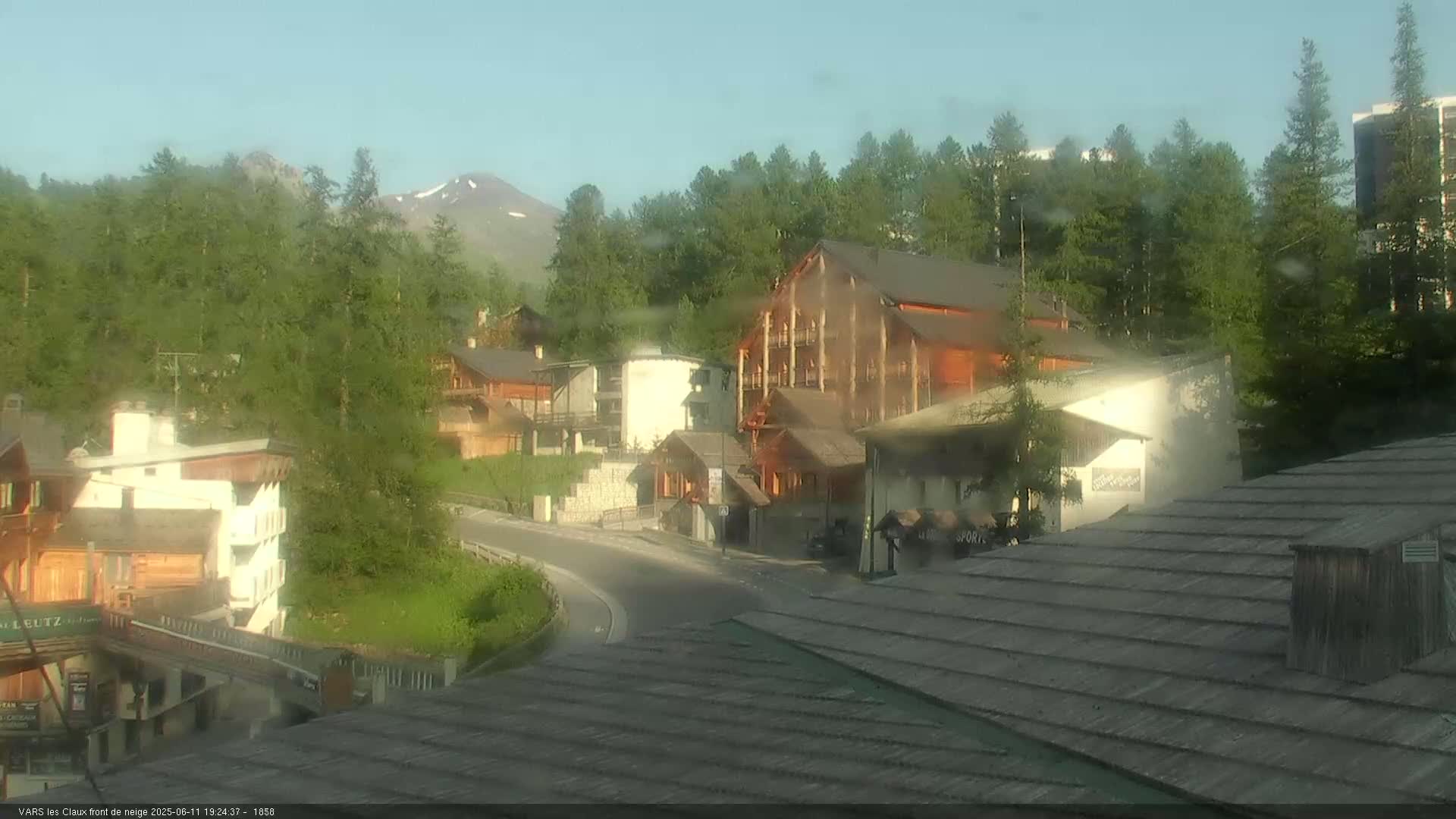 A mountain village nestled among green trees and a clear sky, with a road curving through the buildings.