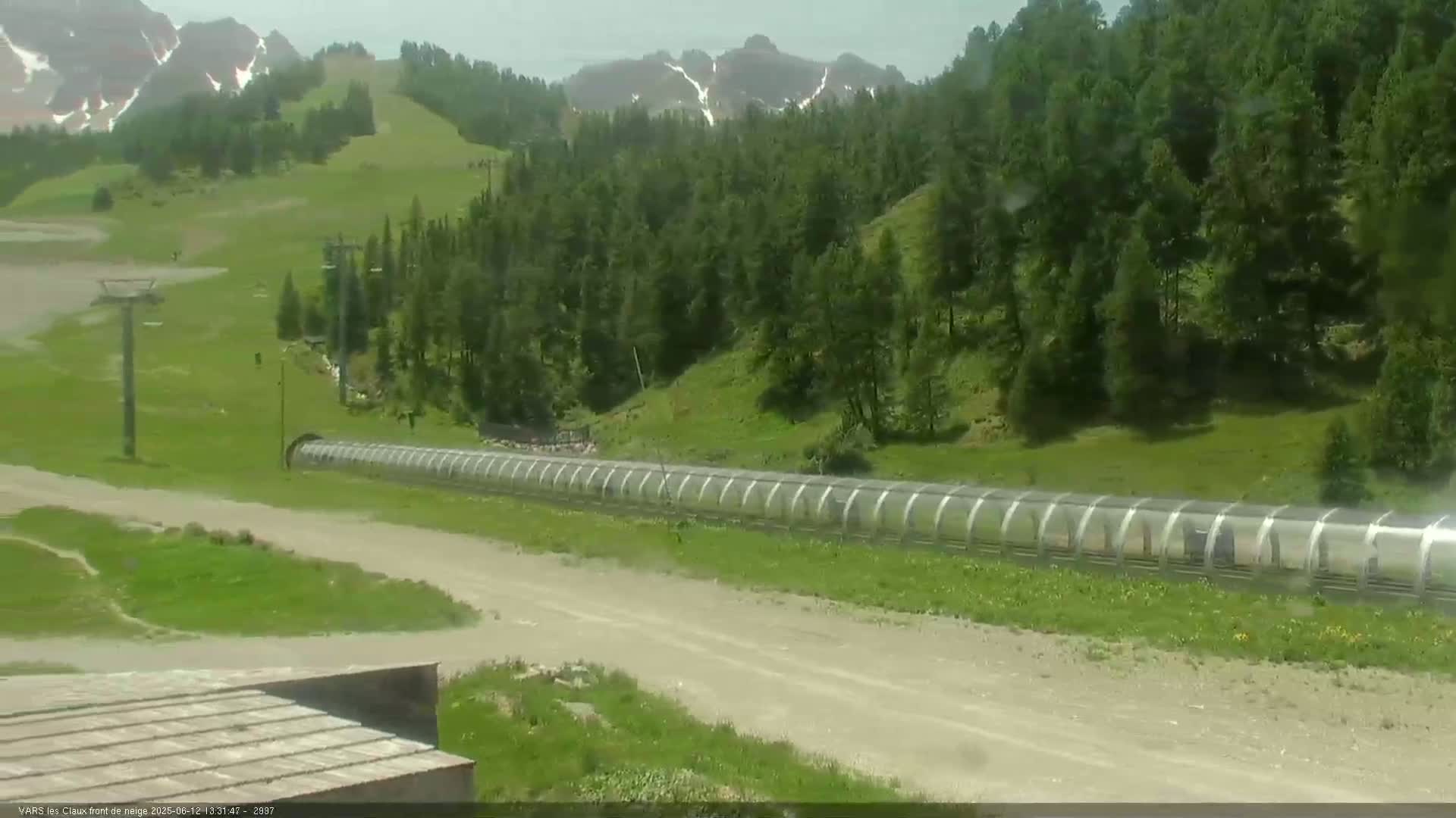 A long, covered structure stretches across a grassy area below a hillside dotted with evergreen trees and mountains in the distance on a partly cloudy day.