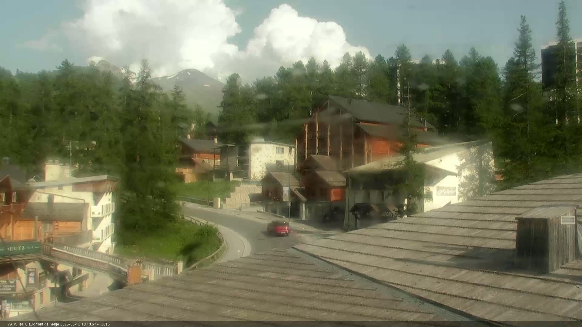 A mountain village with wooden buildings and a red car on a road is seen through rain-streaked glass, with partly cloudy skies and mountains in the background.