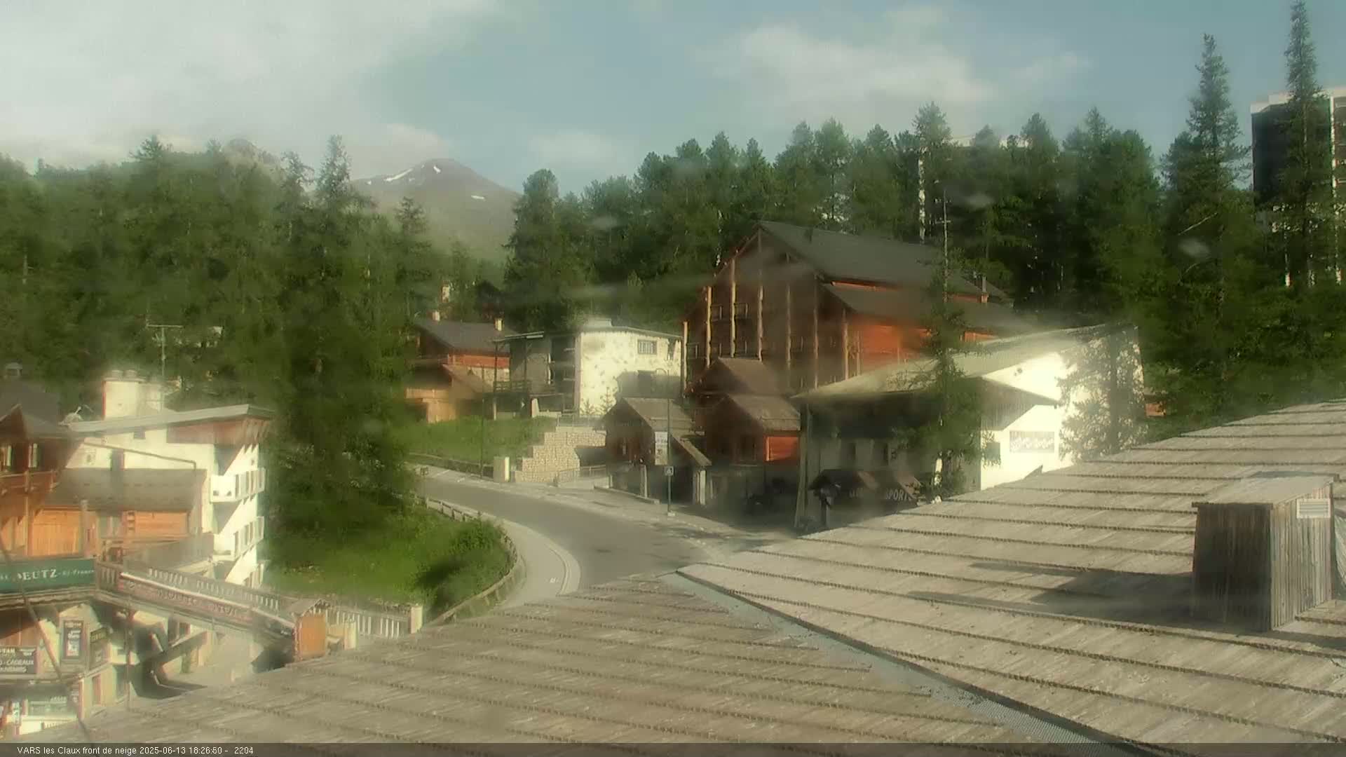 A lightly overcast view of a mountain village with several buildings, a curving road, and green trees, showing light rain on the camera lens.