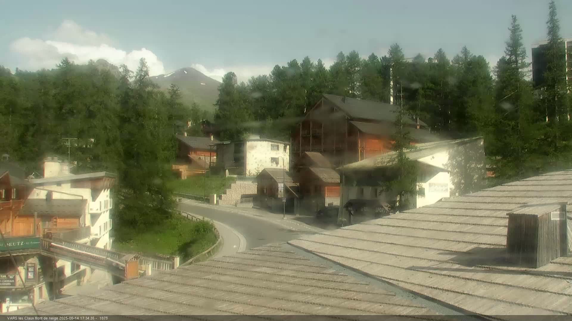 A lightly clouded sky hangs over a mountain village of low buildings and winding roads, with light rain obscuring the view.