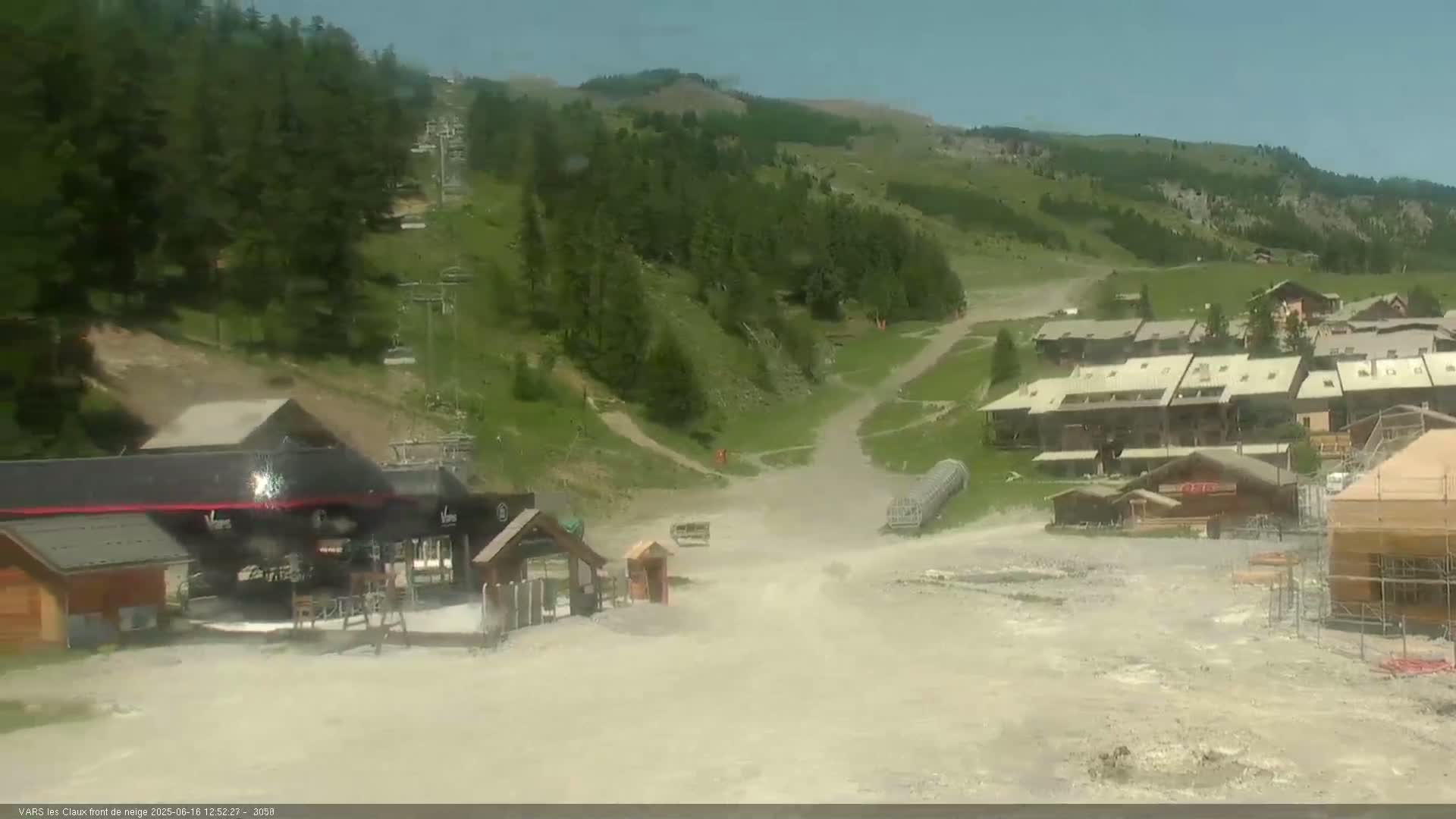 A mostly sunny view of a mountain resort area with buildings, ski lifts, and dry, dusty ground.