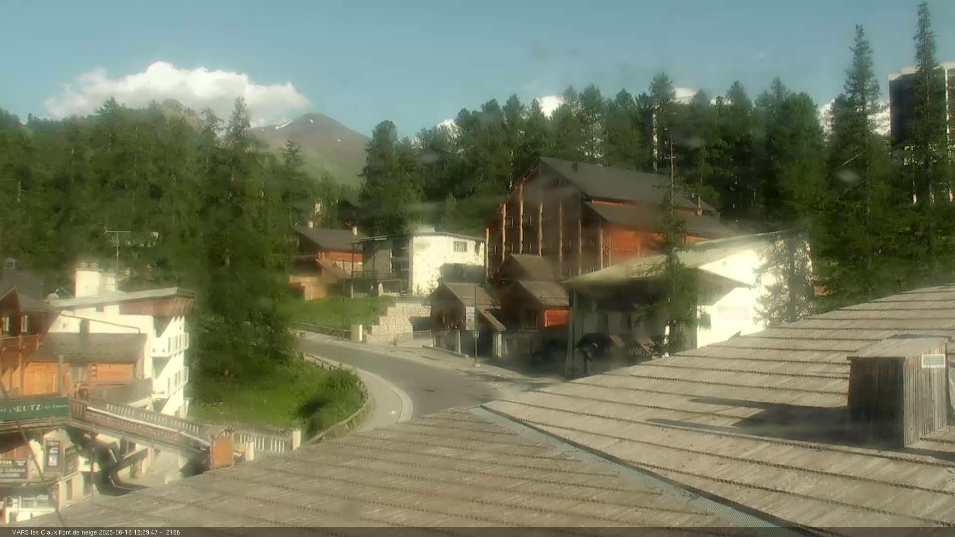 A mountain village with several buildings nestled amongst green trees under a partly cloudy sky.