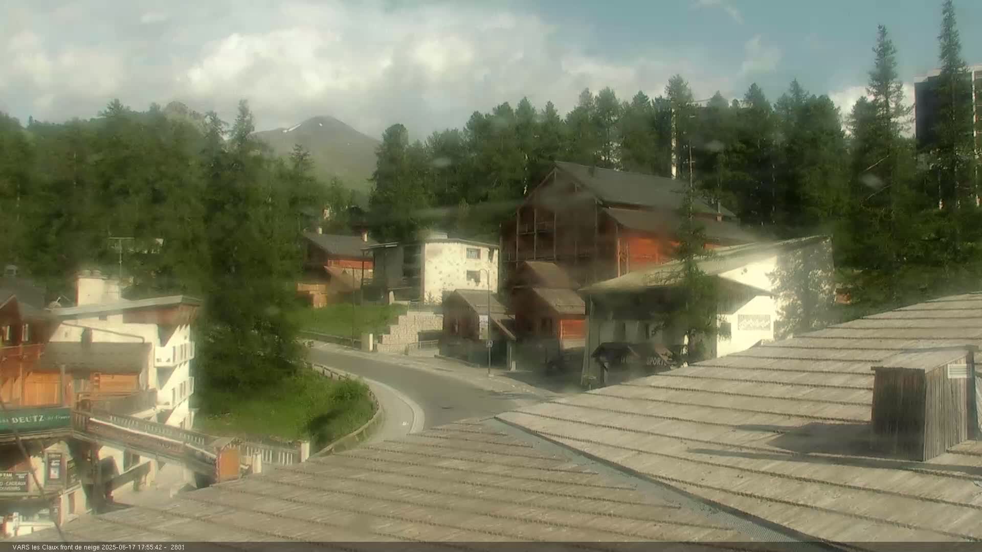 A partly cloudy day reveals a mountain village nestled amongst a dense forest, with a road curving through the buildings.