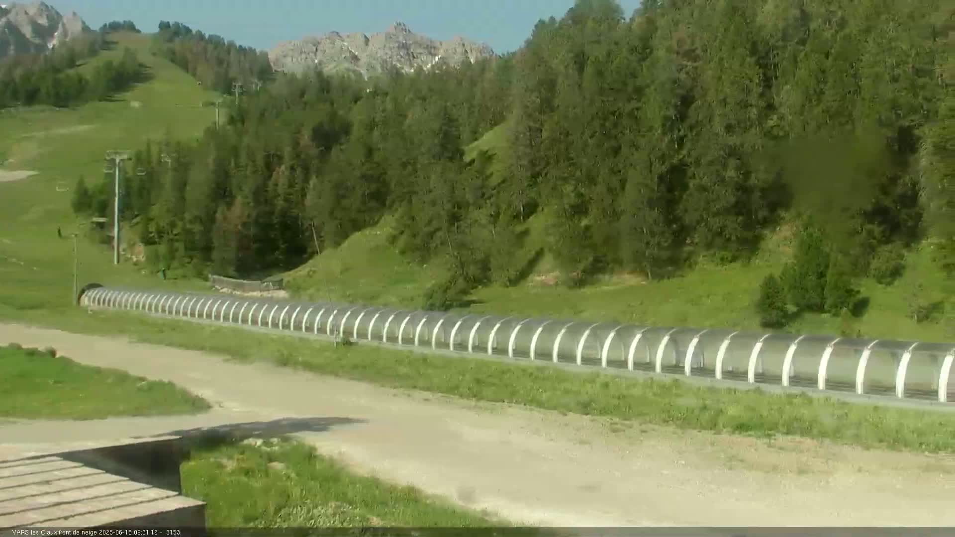A long, covered structure runs horizontally across a grassy hillside, with a dirt road leading to it, under a sunny sky and surrounded by green vegetation and mountains in the background.