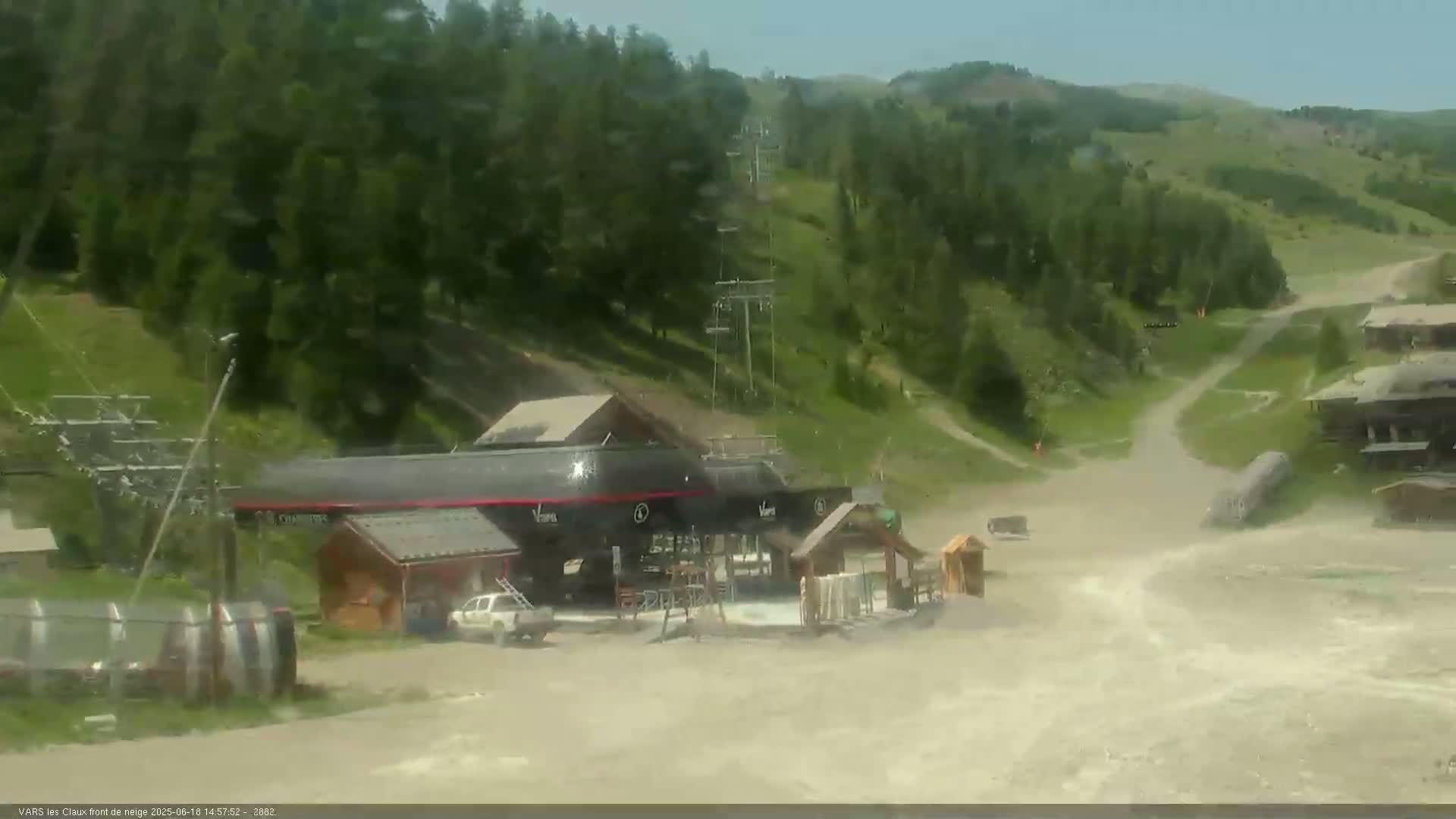 A ski lift base area with several buildings and a dirt road, surrounded by green hills under a clear sky.