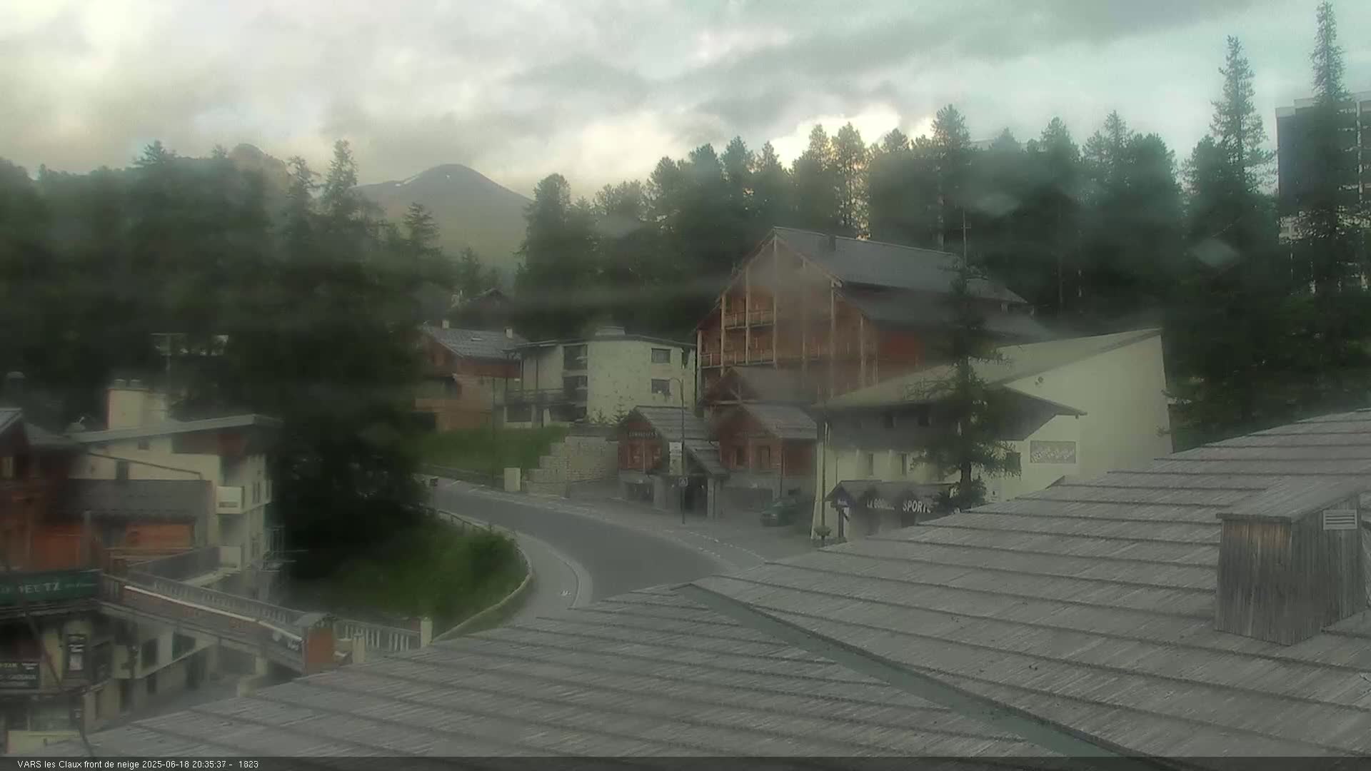 A mountain village with several buildings is seen under a cloudy, rainy sky, with mountains visible in the background.