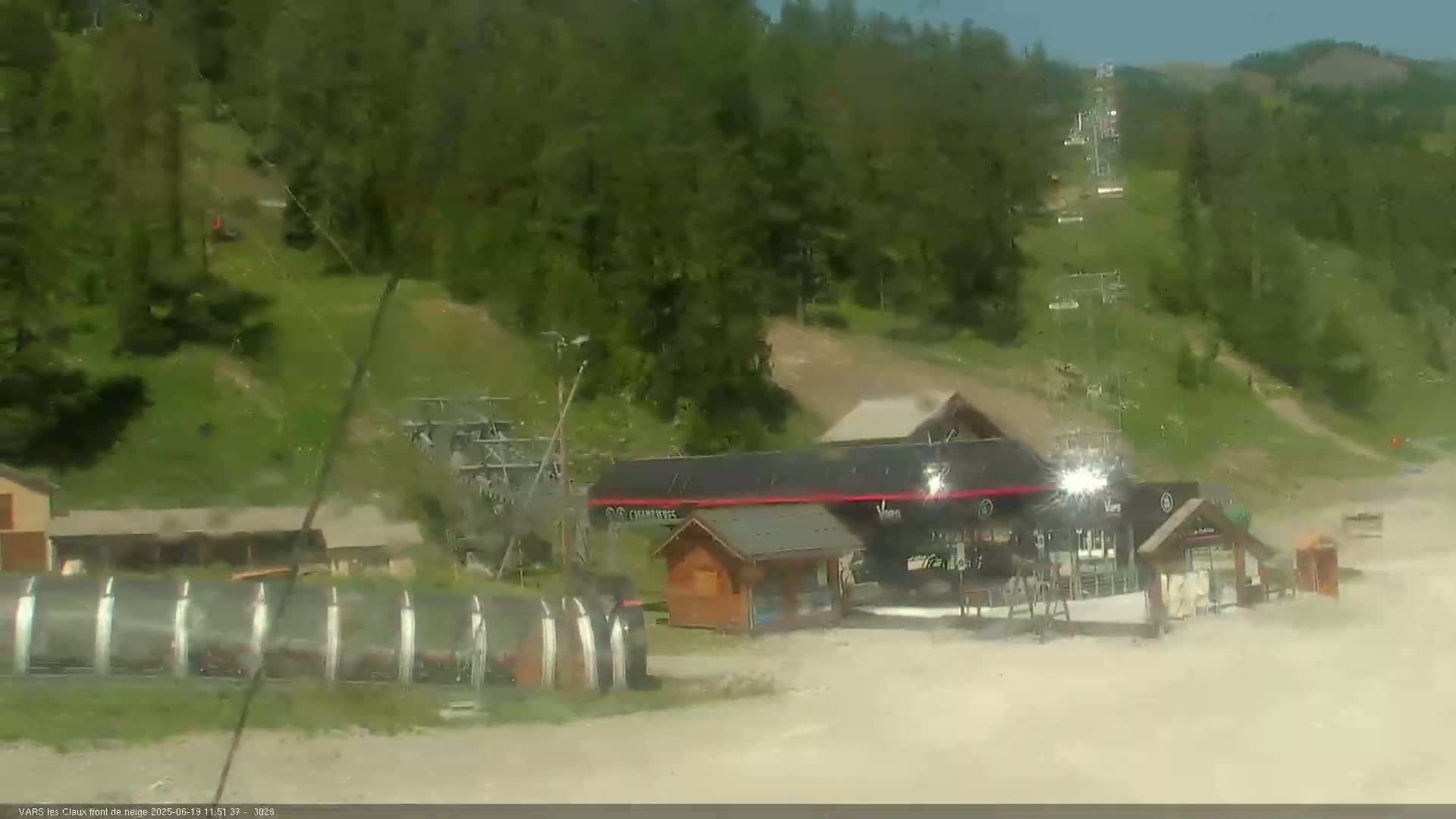 A ski resort base area with a chairlift, several buildings, and a long, covered structure is visible on a sunny day.