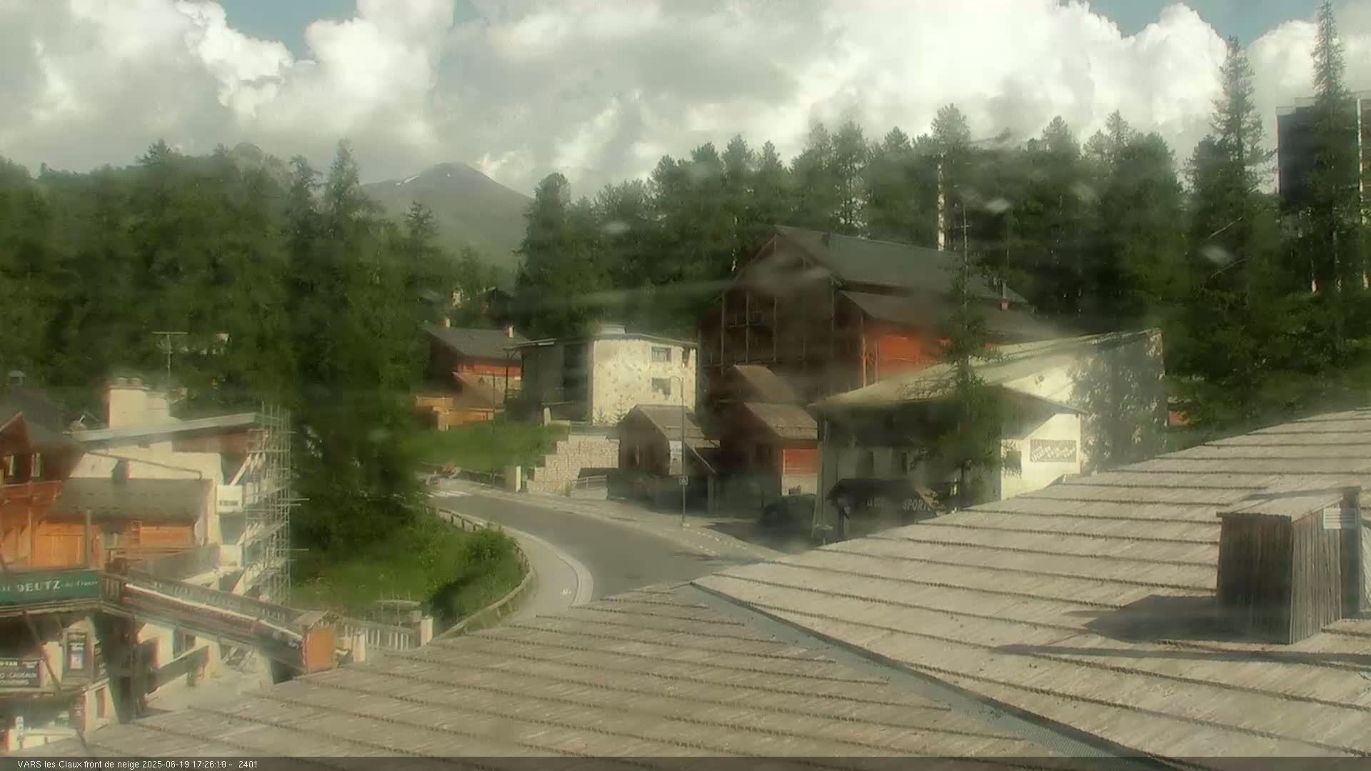 A mountain village with several buildings, some under construction, is visible beneath a partly cloudy sky, with rain obscuring the view.