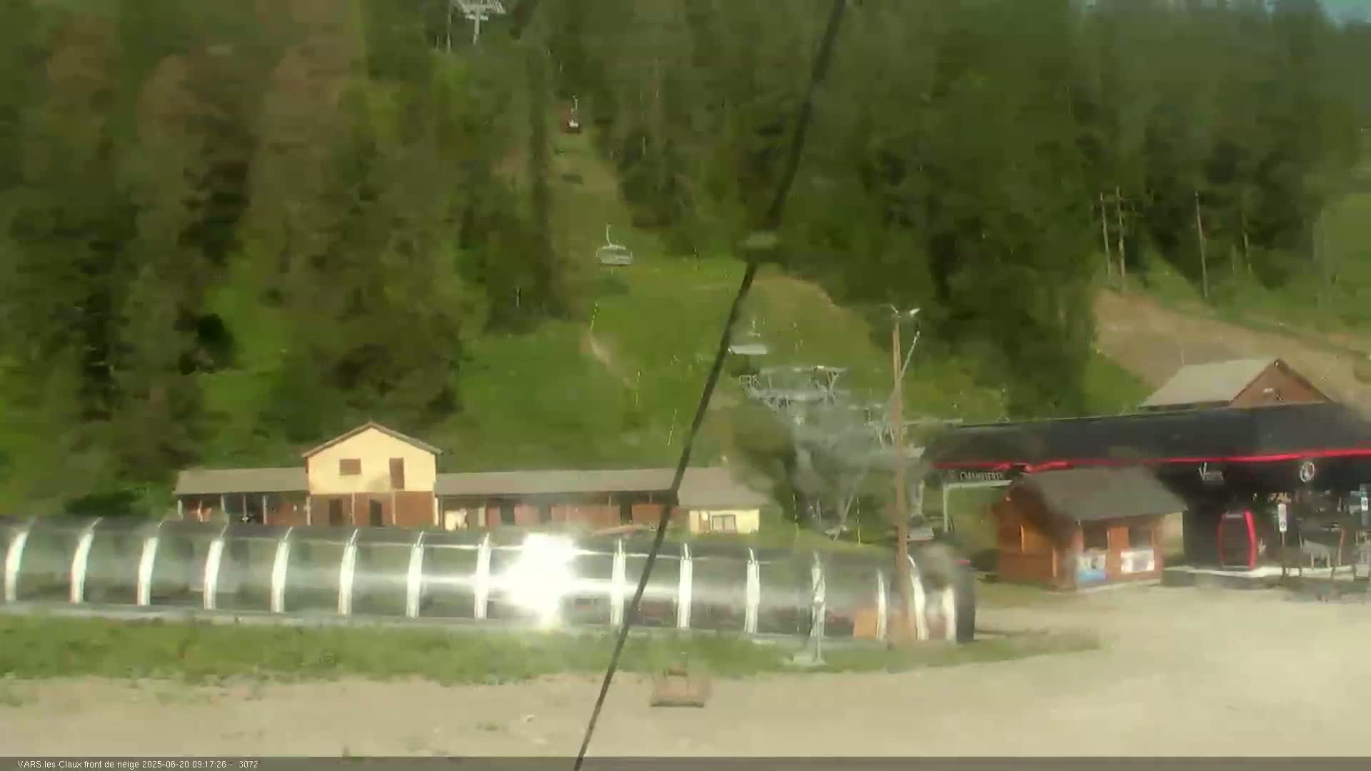 A ski lift and several buildings are situated on a hillside covered in lush green trees under a mostly clear sky.