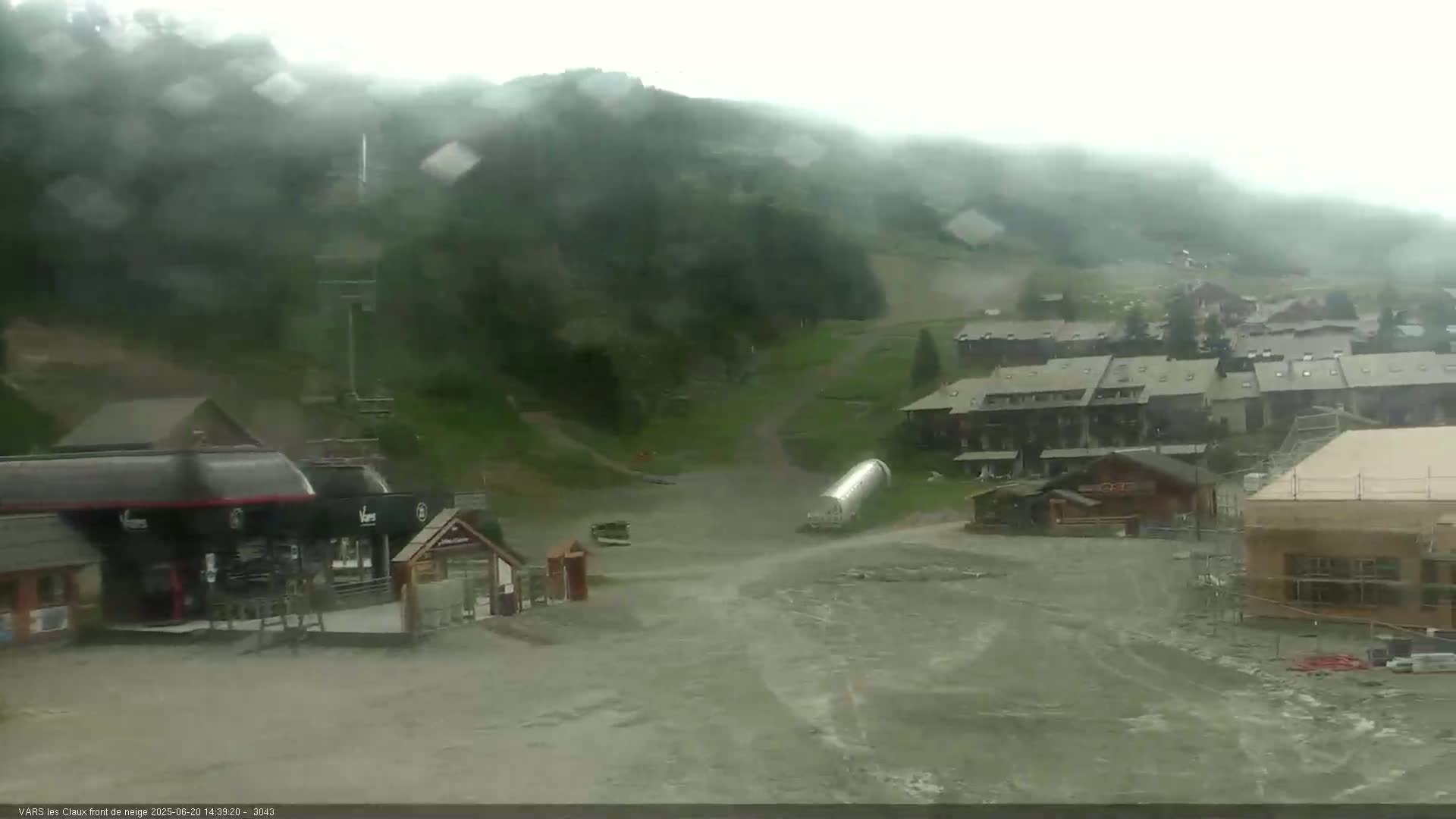 A rain-soaked mountain village nestled in a valley, with buildings and construction sites visible under a misty sky.