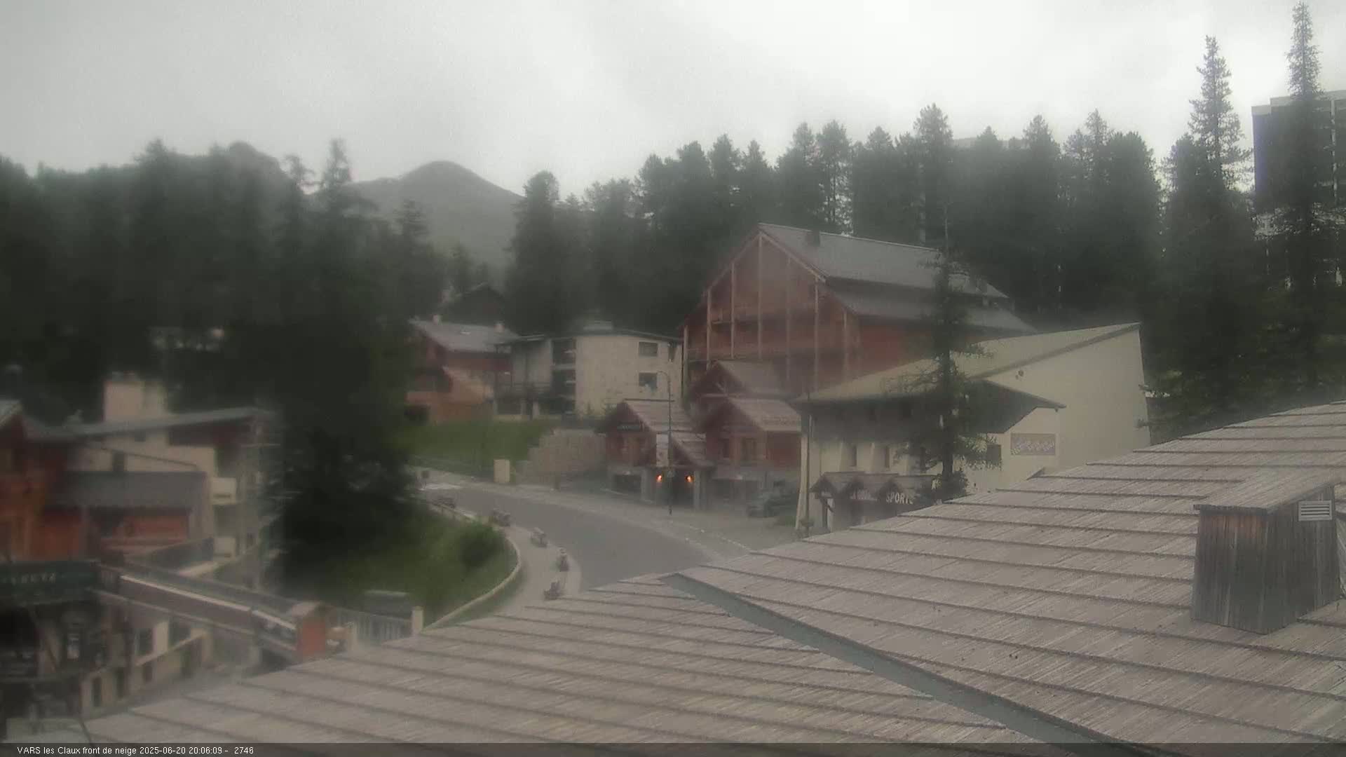 A foggy mountain village scene shows several chalet-style buildings and a curving road, with overcast skies.