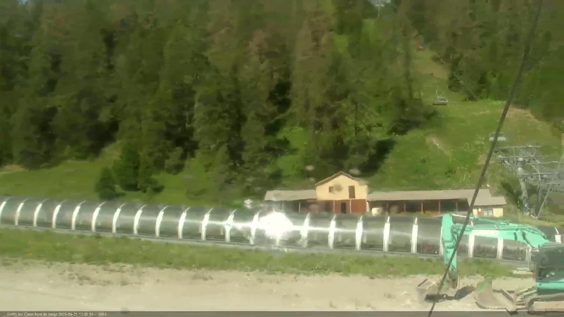 A long, transparent covered structure is situated near a small building amidst a green landscape under sunny conditions, with a teal excavator visible at the structure's end.