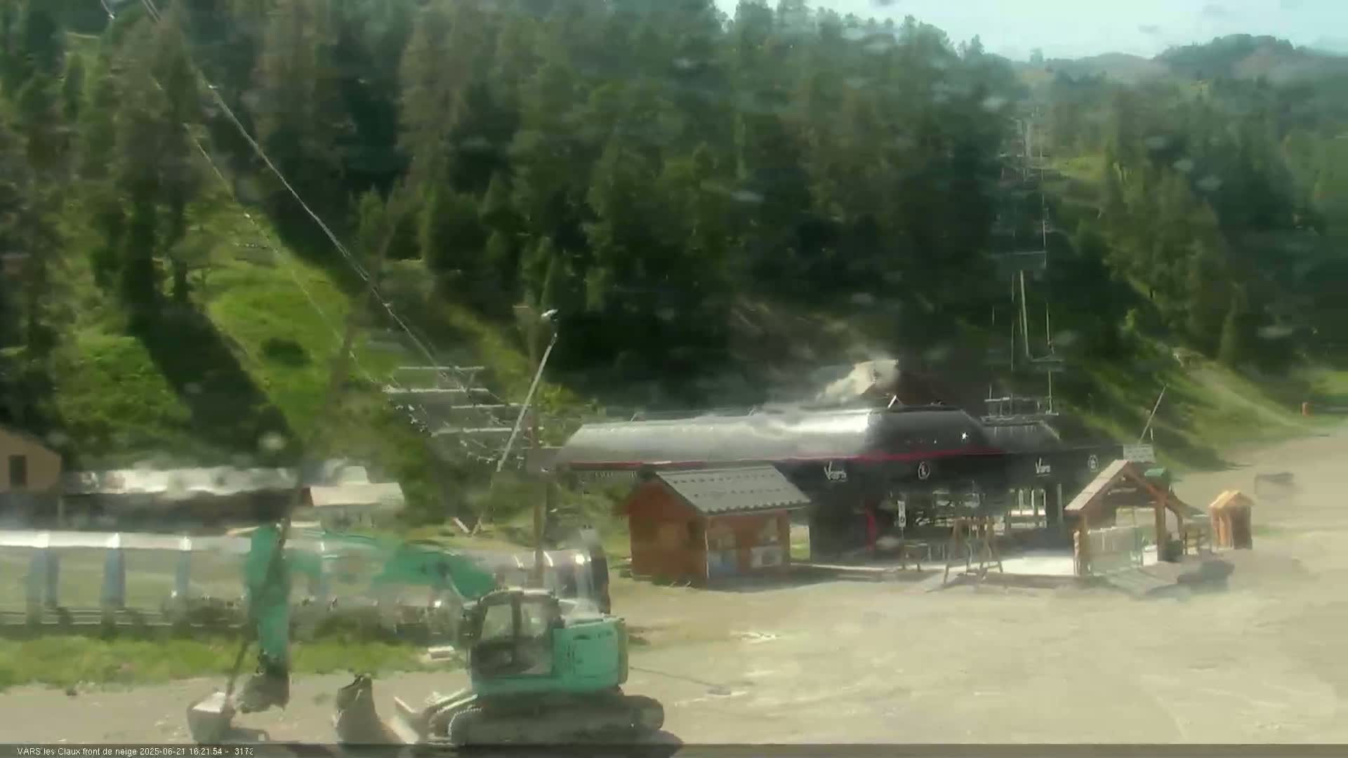A teal excavator sits near a ski lift base station in a mountainous area on a partly cloudy day.