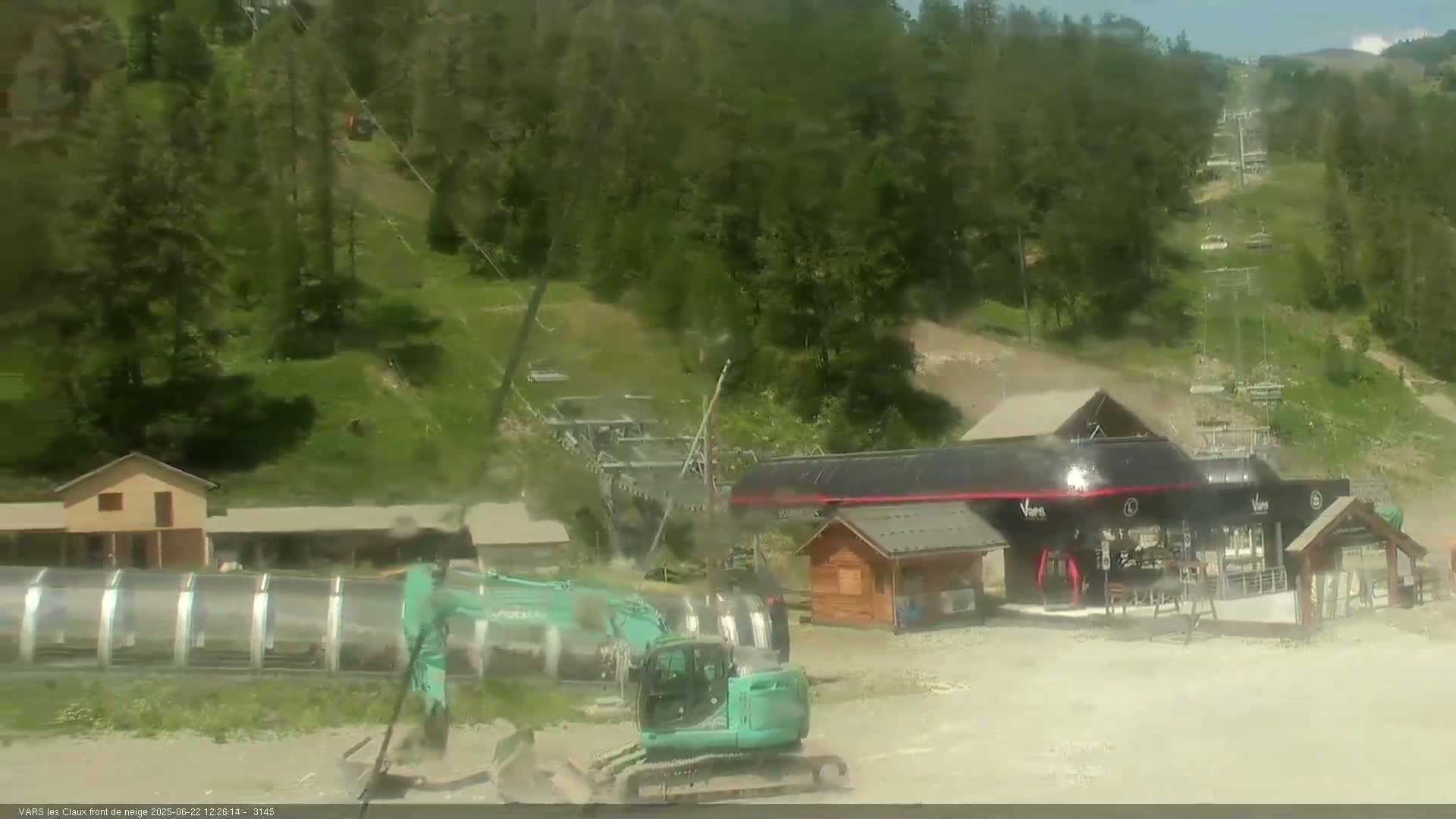 A green excavator sits near a ski lift base station on a sunny day amidst a verdant, hilly landscape.