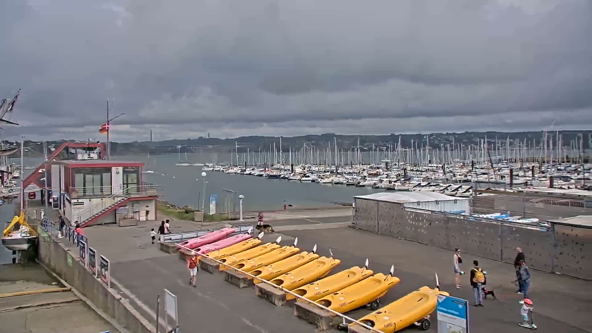 A marina with numerous sailboats in the water under a cloudy sky, kayaks lined up on the shore near a small building, and several people walking nearby.