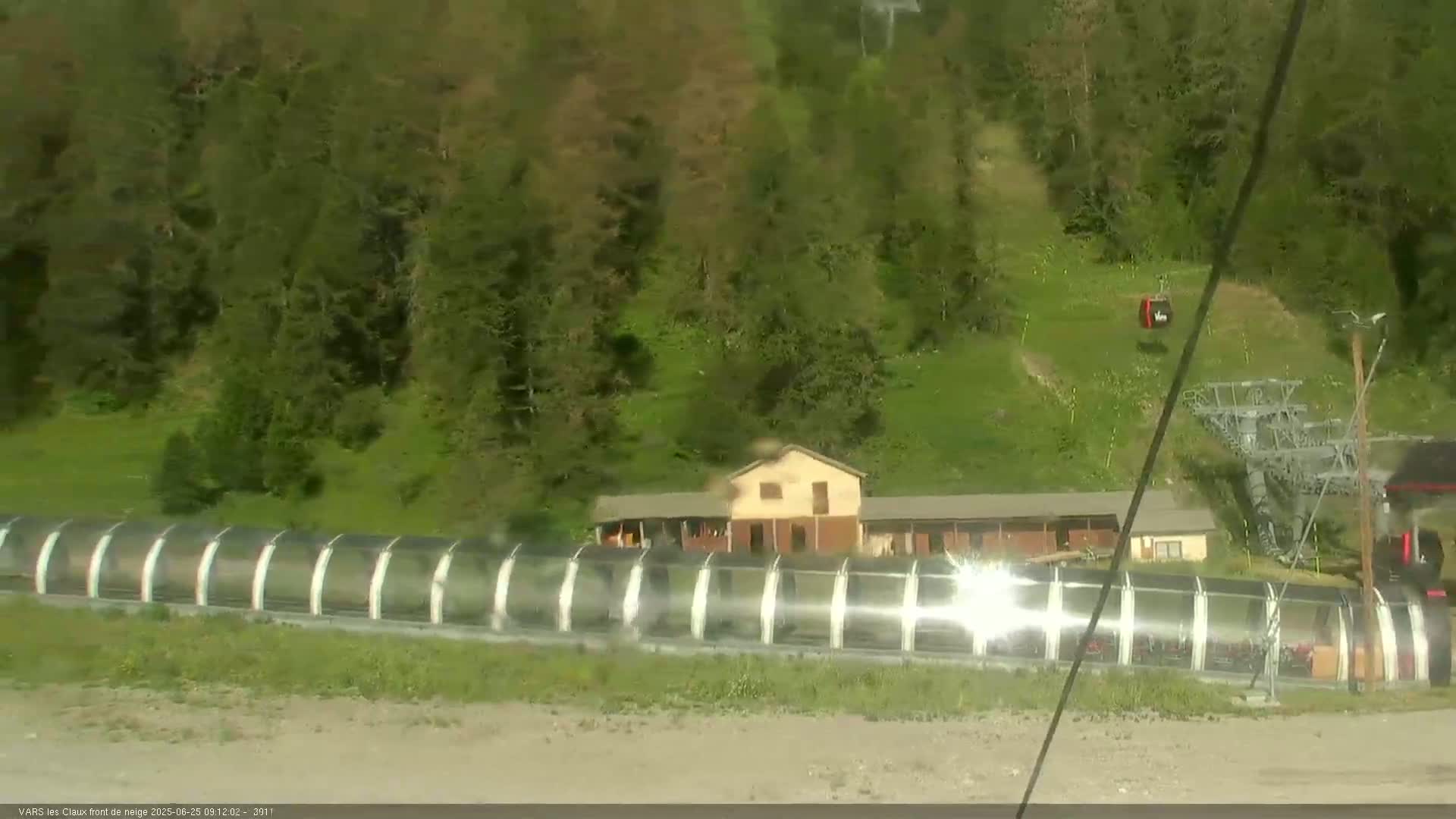A long, covered walkway sits below a building in a mountainous area with green grass and trees, under a sunny sky.