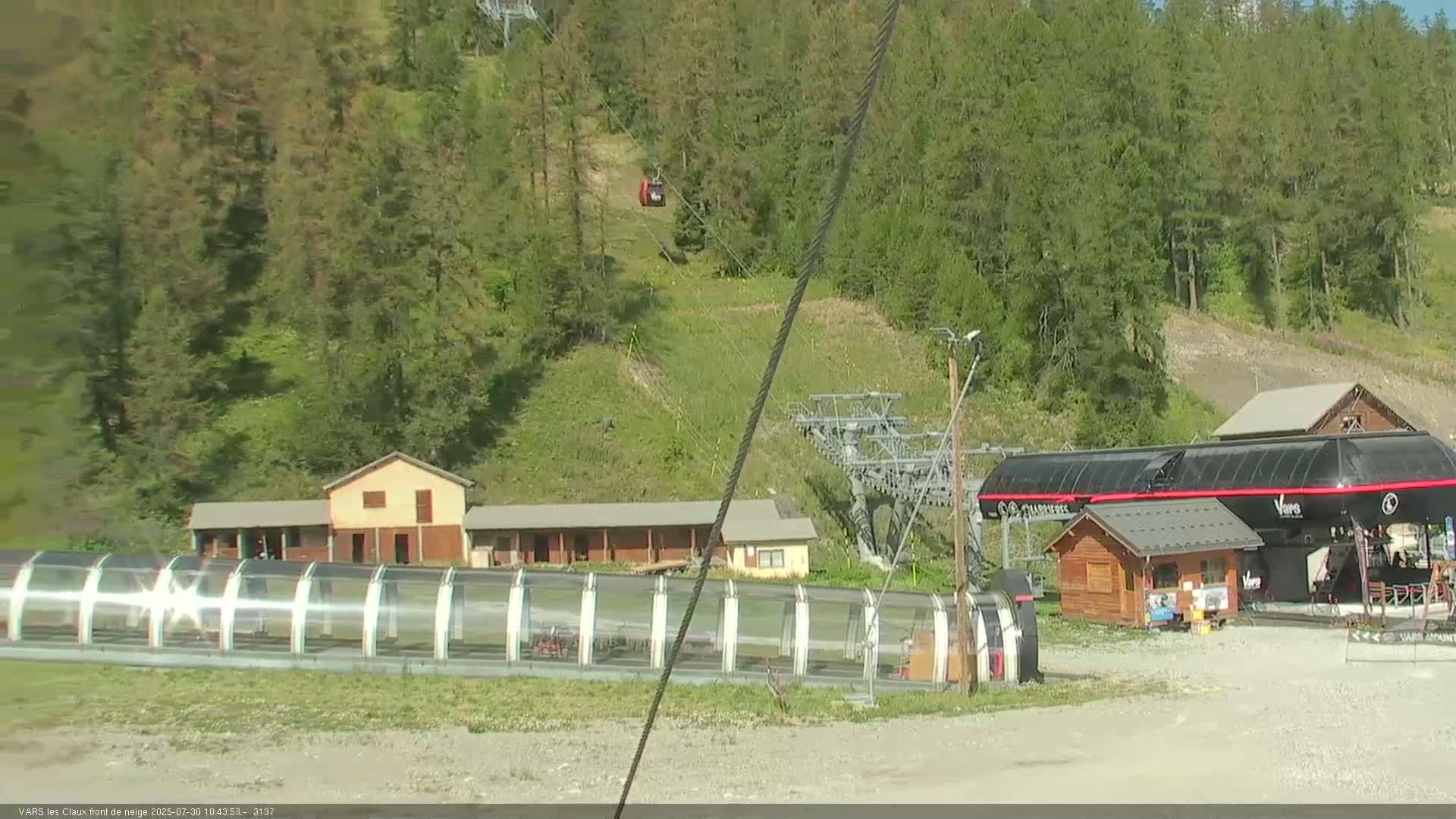 A ski lift station with buildings and a covered area sits at the base of a hill covered in lush green trees under a sunny sky.