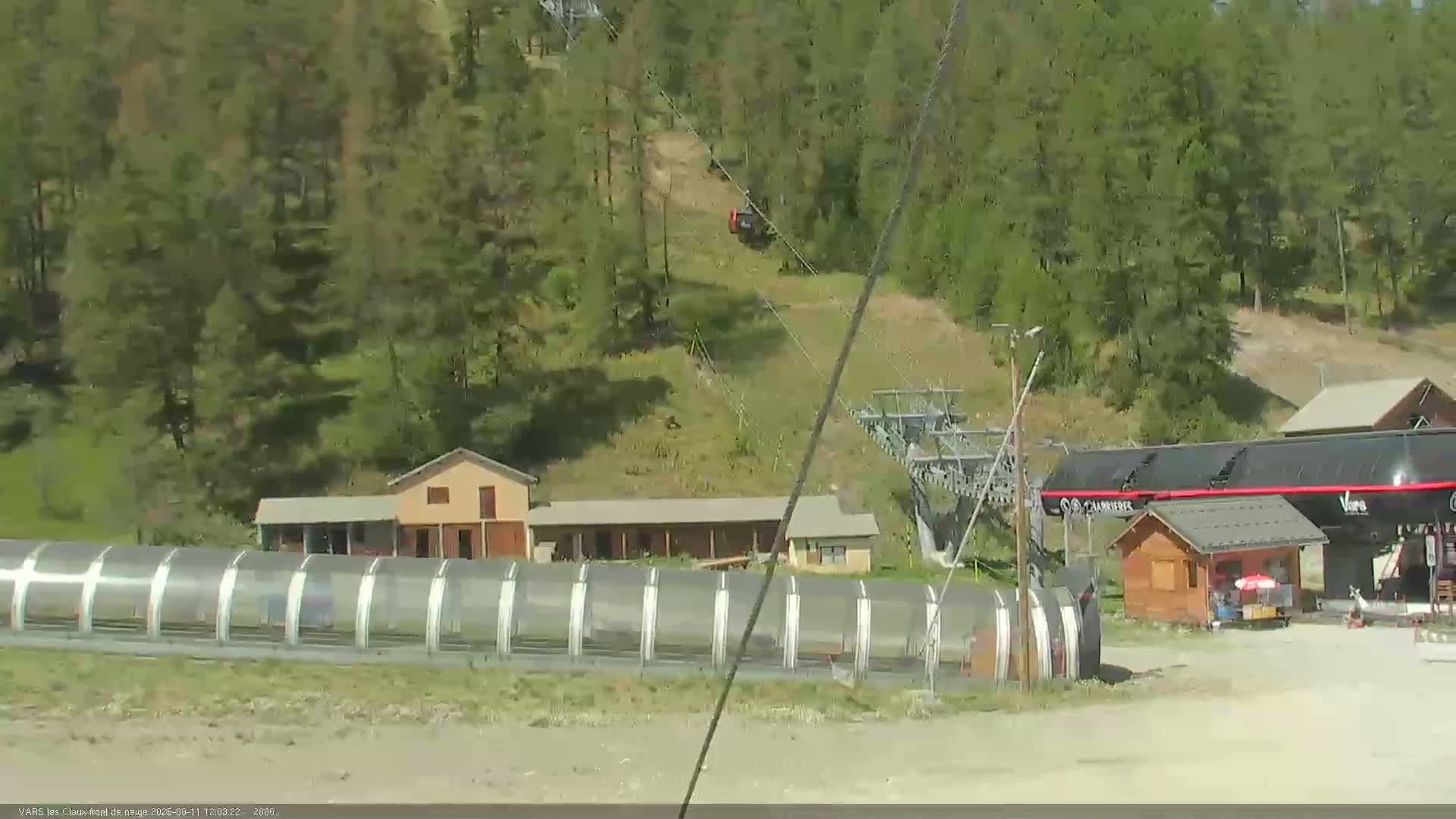 A chairlift ascends a hill covered in green trees on a sunny day, passing over a few small buildings and a long, transparent structure.