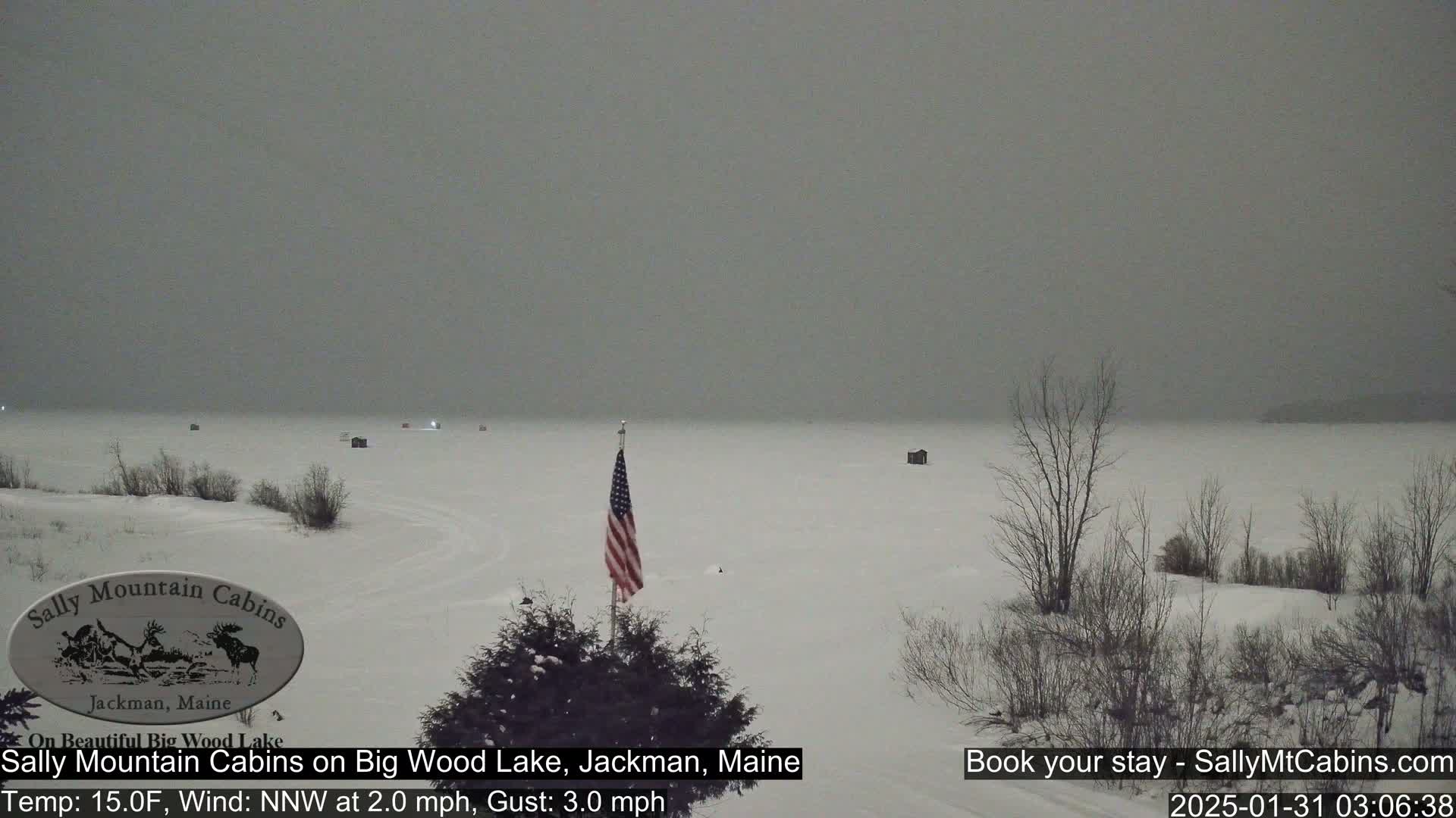 A snow-covered frozen lake is seen under an overcast sky, with a few small structures visible on the ice and a US flag standing near the foreground.