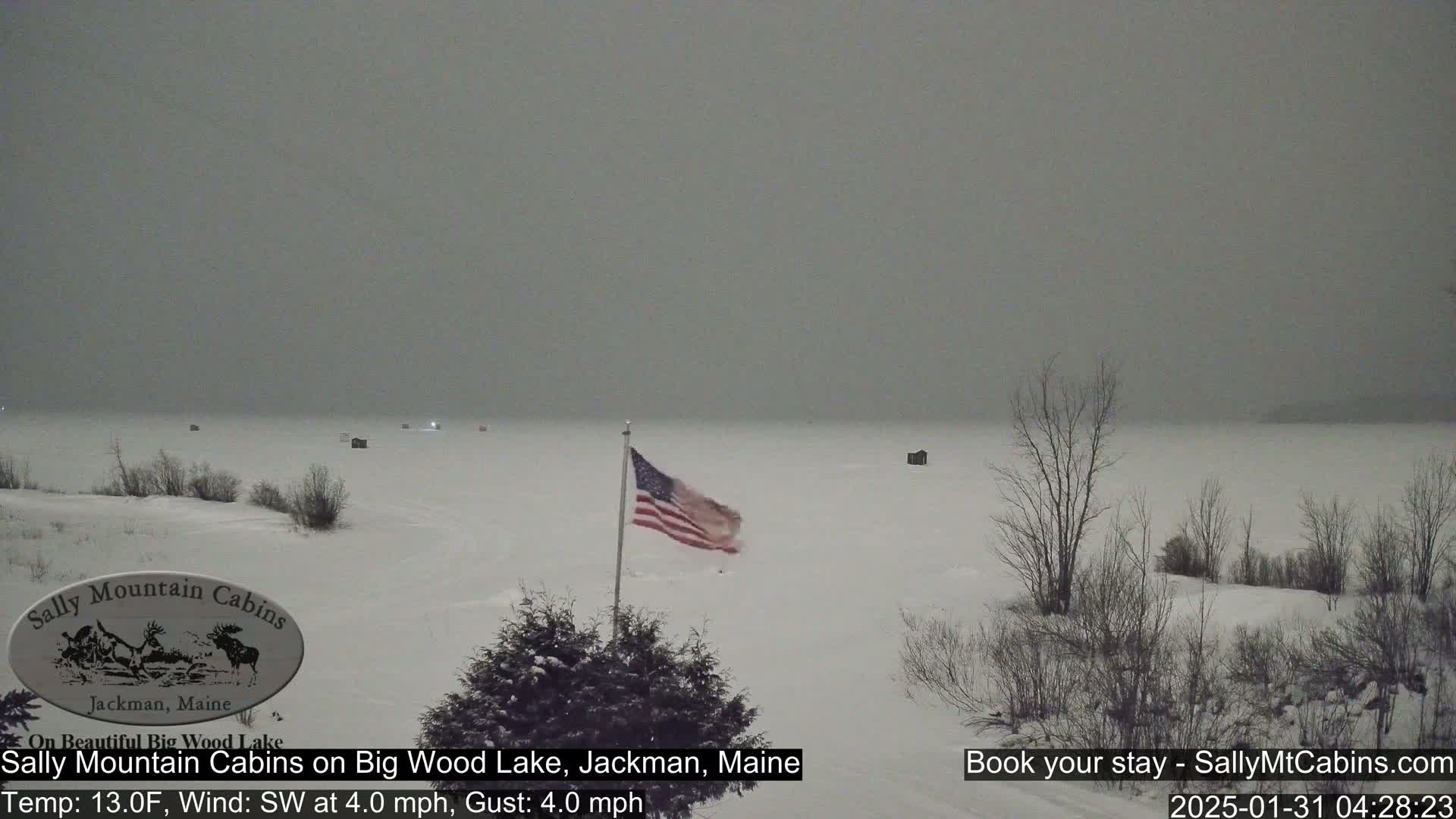 A snow-covered, frozen lake is visible, with several small structures on the ice, a US flag waving in the light snowfall, and bare trees lining the shoreline under a gray sky.