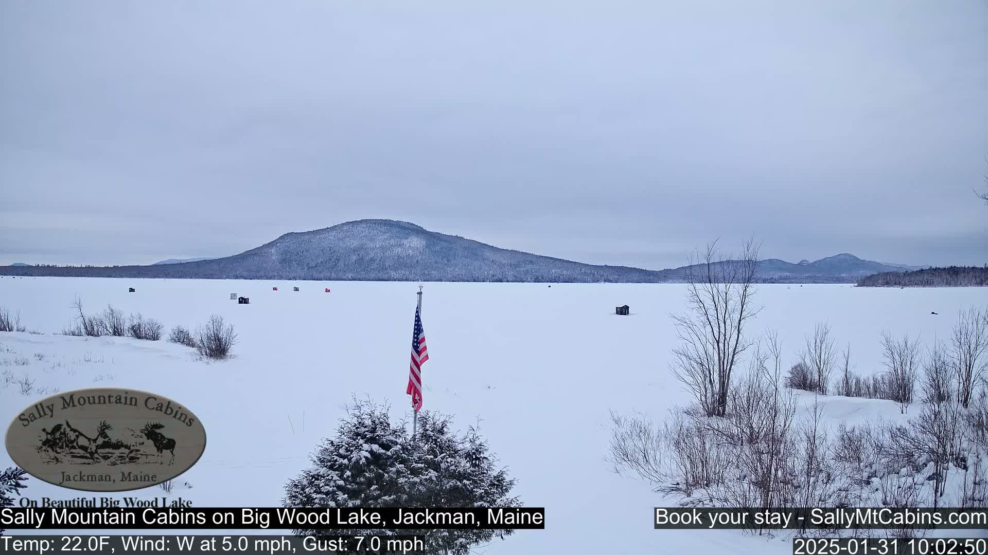 A snow-covered frozen lake stretches to a snow-dusted mountain range under a cloudy sky, with a few ice fishing huts scattered across the ice and an American flag standing in the foreground.