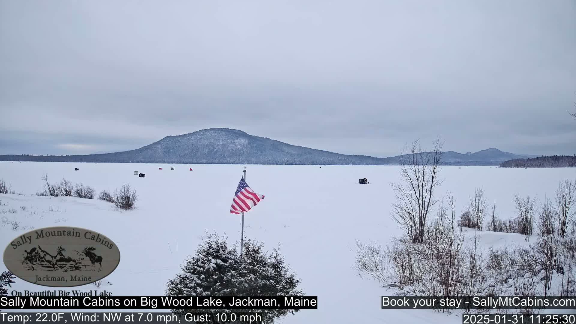 A snow-covered frozen lake stretches to a distant mountain range under a cloudy sky, with a few ice fishing shelters scattered across the ice and an American flag waving in the foreground.