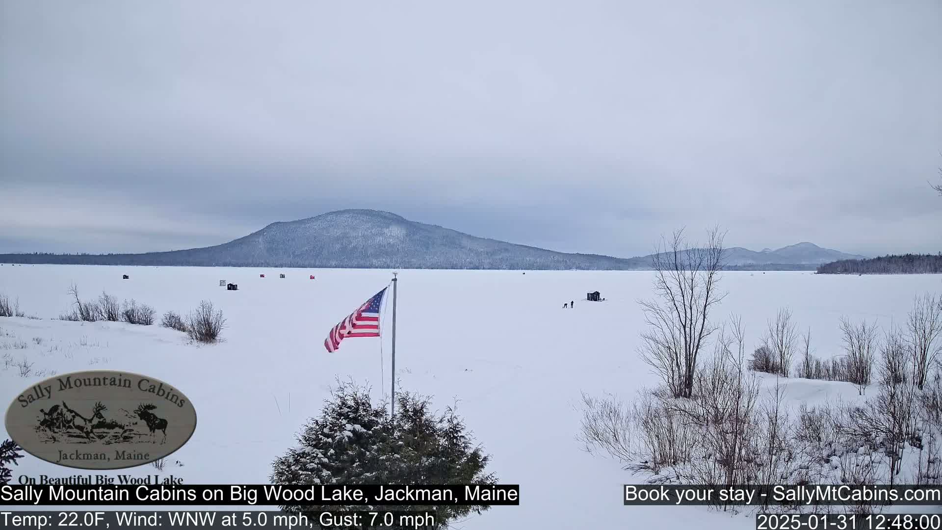 A snow-covered frozen lake stretches to a distant mountain range under an overcast sky, with several ice fishing huts visible and an American flag planted nearby.