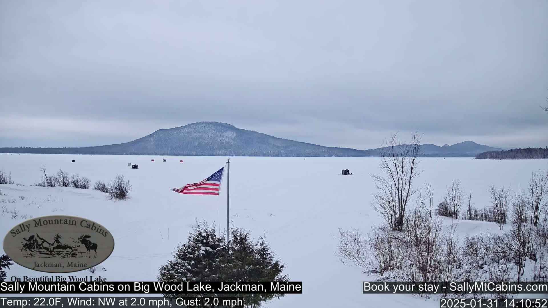 A snow-covered frozen lake stretches to a distant mountain range under an overcast sky, with a few ice fishing shacks and an American flag visible.