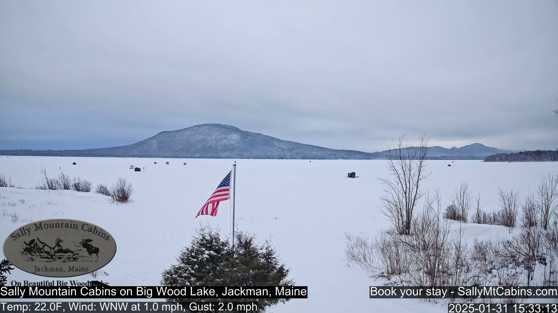 A snow-covered frozen lake stretches to a distant mountain range under a cloudy sky, with a few ice fishing shanties scattered across the ice and an American flag planted near the foreground.