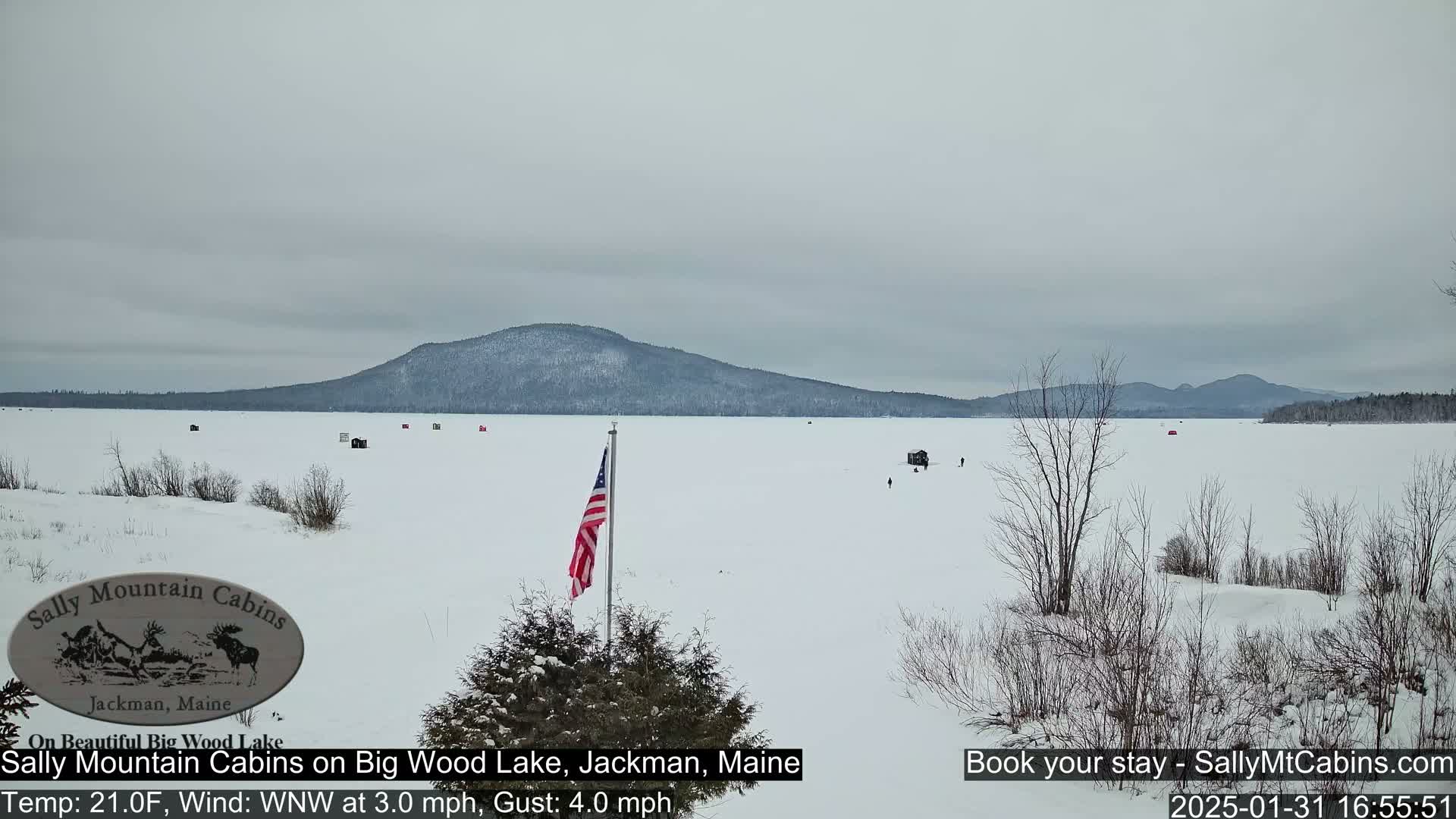 A frozen lake stretches to a snow-covered mountain range under an overcast sky, with several ice fishing shelters scattered across the ice and an American flag in the foreground.